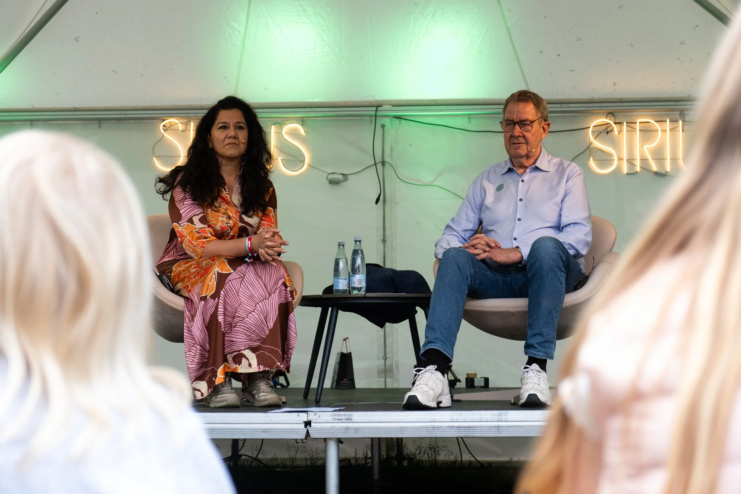 To personer sidder på en scene og deltager i en samtale foran et publikum. Baggrund med neonlys, der siger 'SIRIUS'.