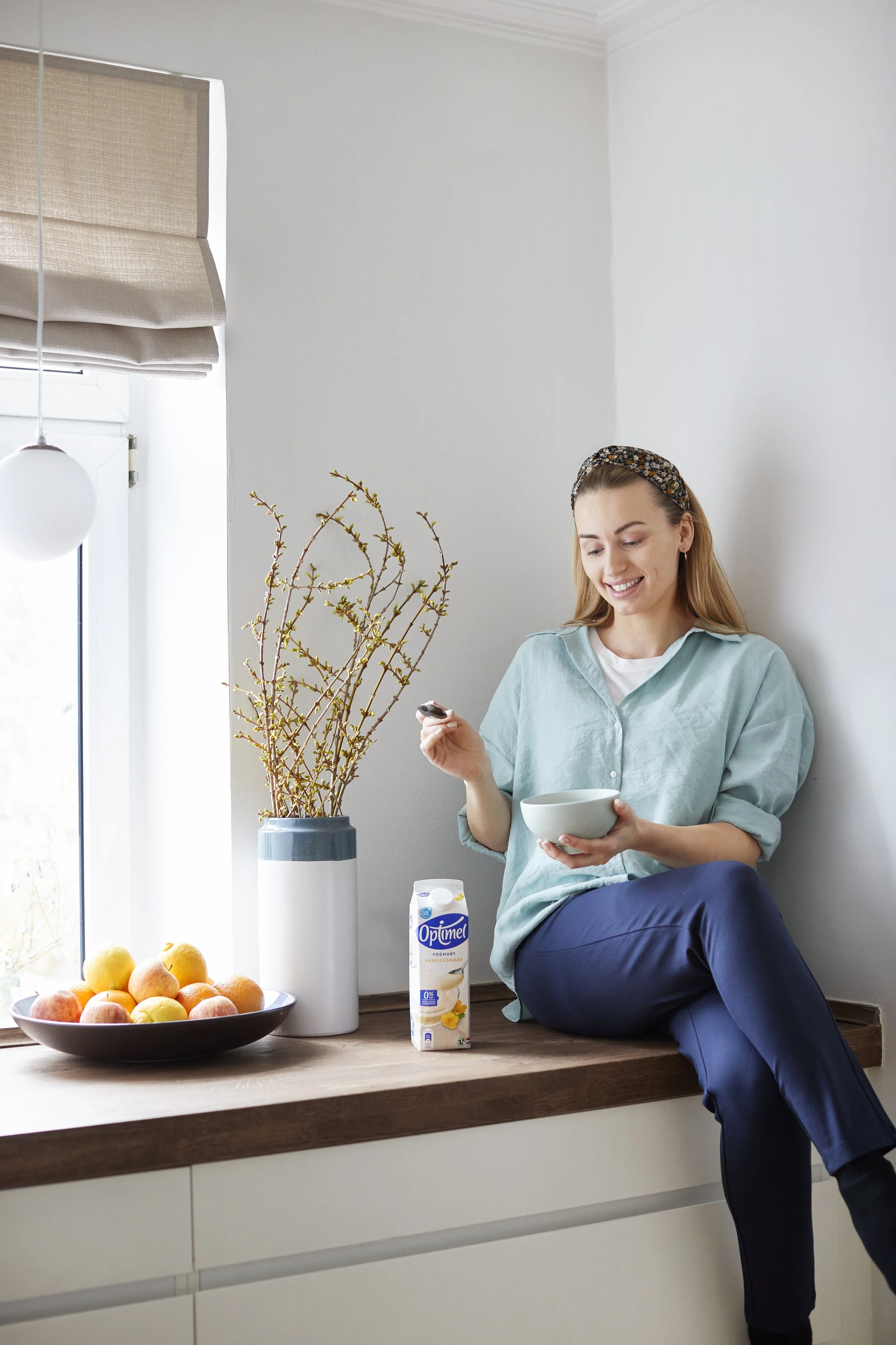 Kvinde sidder på køkkenbord, spiser morgenmad, med appelsiner og en mælkekarton på bordet.