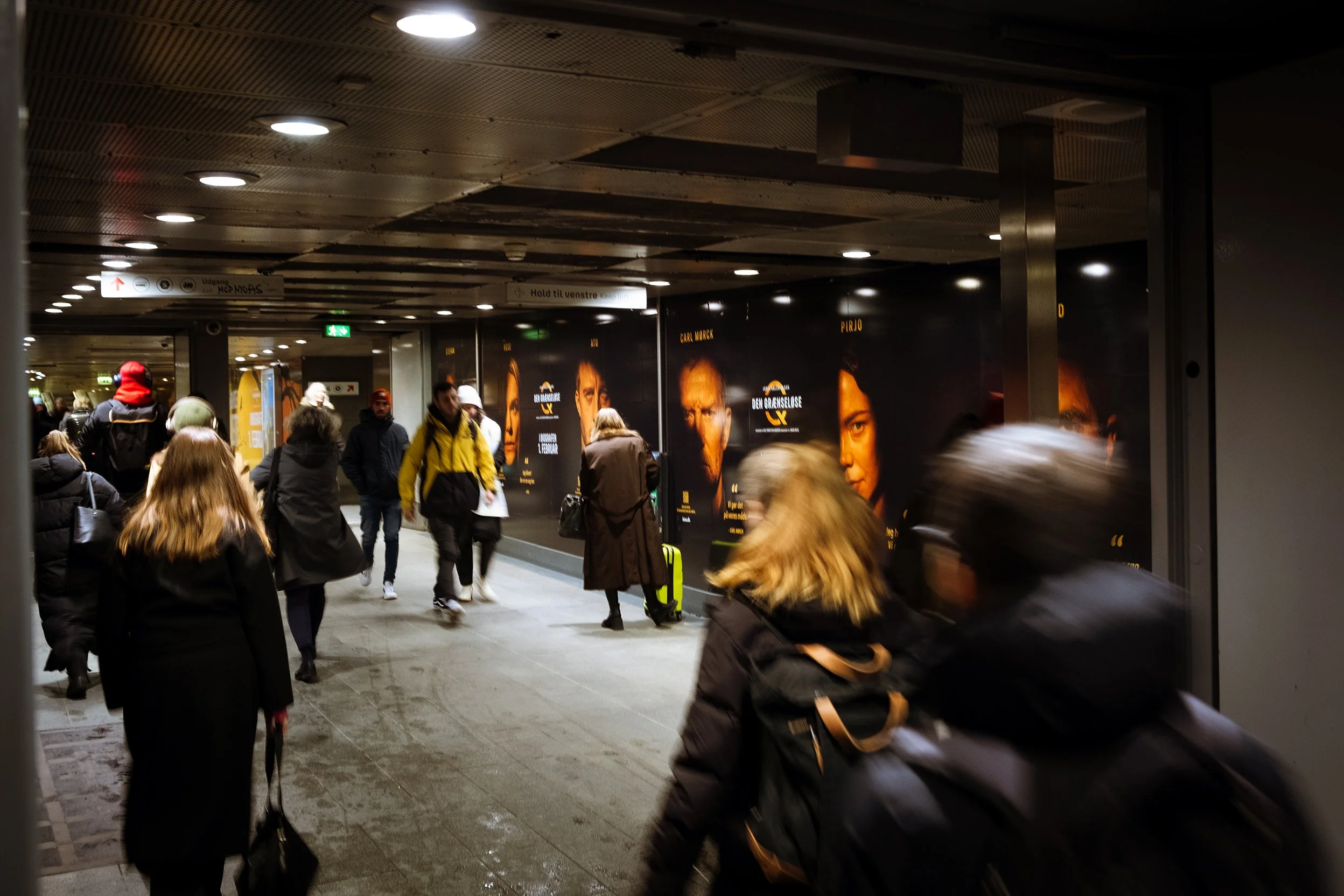 Folk går gennem en undergrundsstation med reklamer og skilte, nogle trækker kufferter.