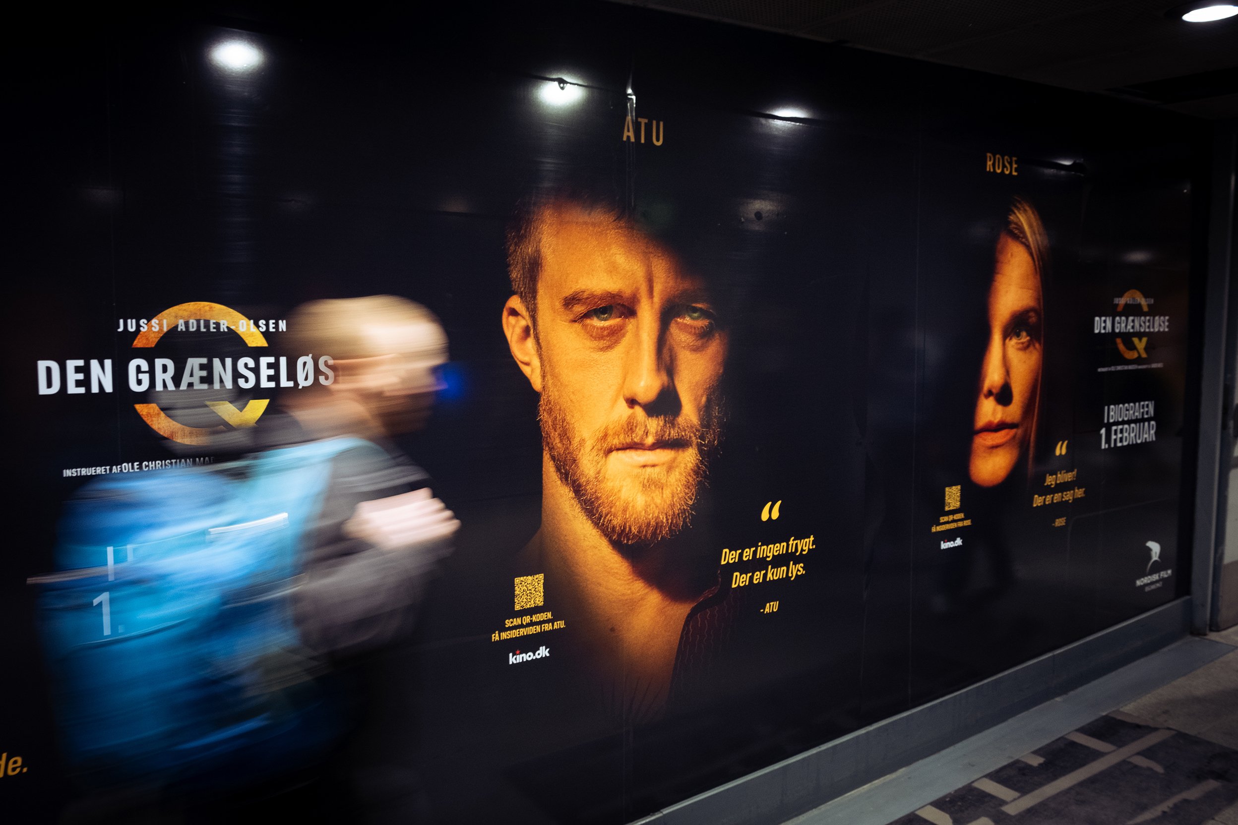 En hurtigt bevægende person passerer foran en sort væg med plakat for filmen "Den Grænseløs", der viser to skuespillere, en mand og en kvinde, med tekst på dansk.