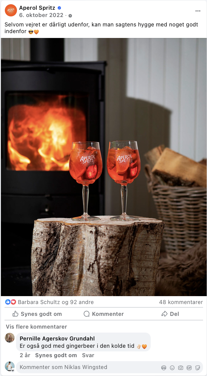 To Aperol Spritz drinks med is på en trætønde, foran en brændeovn med flammer, i en hyggelig indendørs setting.