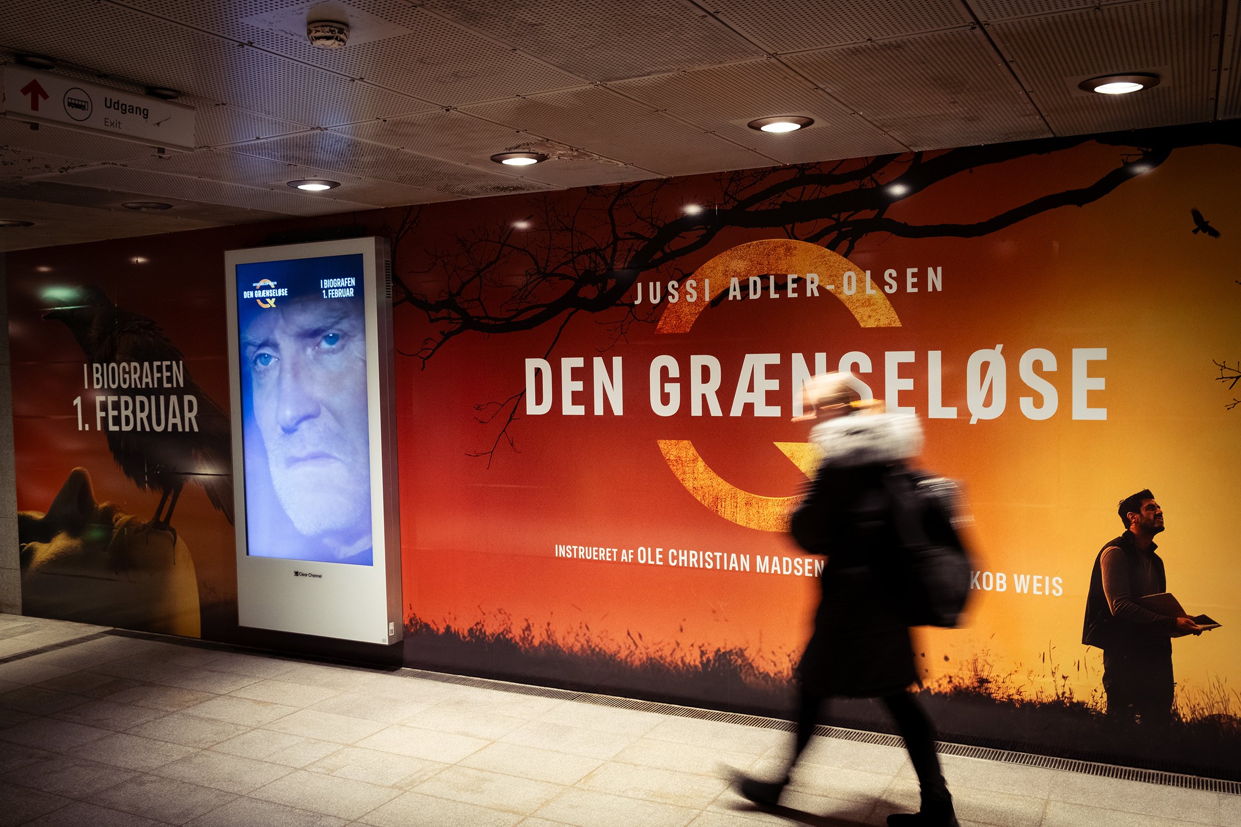Kunstnertekst og plakater for filmen 'Den Grænseløse' med Jussi Adler-Olsen, vist på en væg i en undergrundsstation, med en person gående forbi.