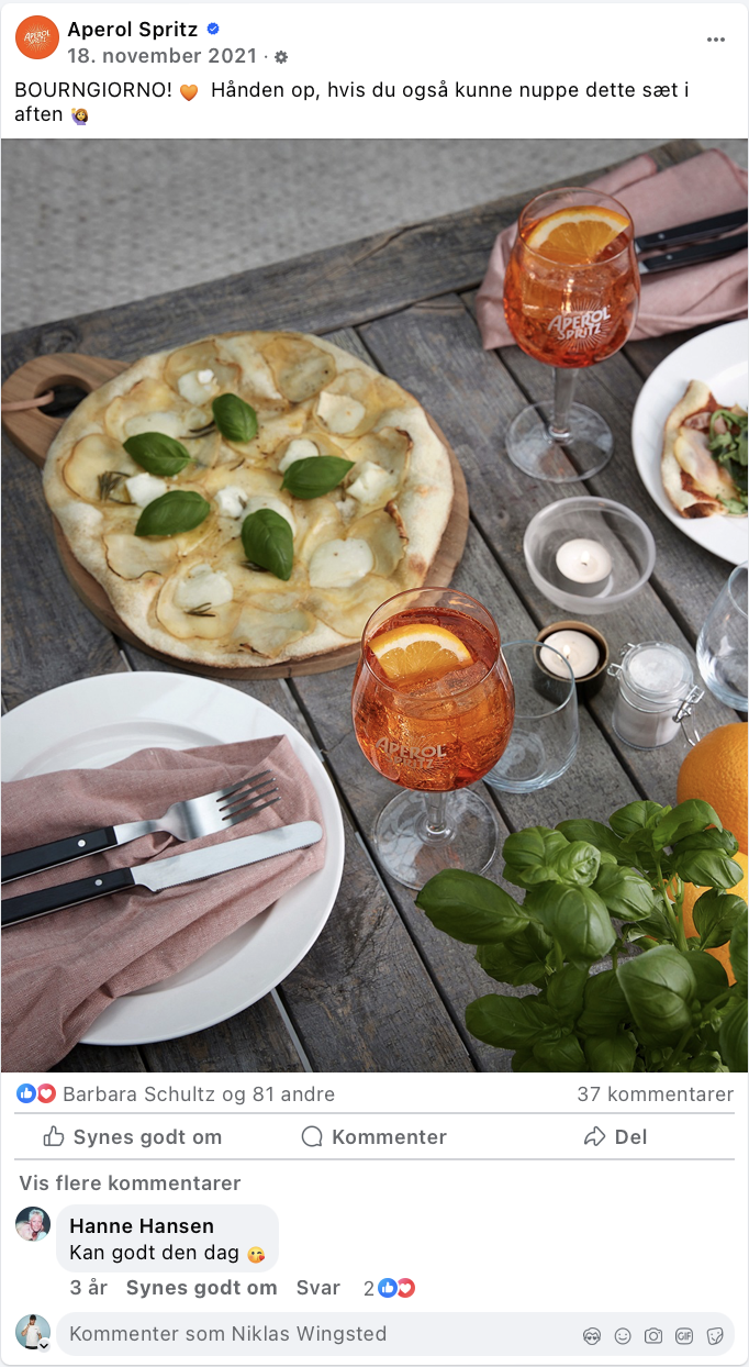 Et bord med pizza, to glas Aperol Spritz, en tallerken med mad, og en grøn plante.