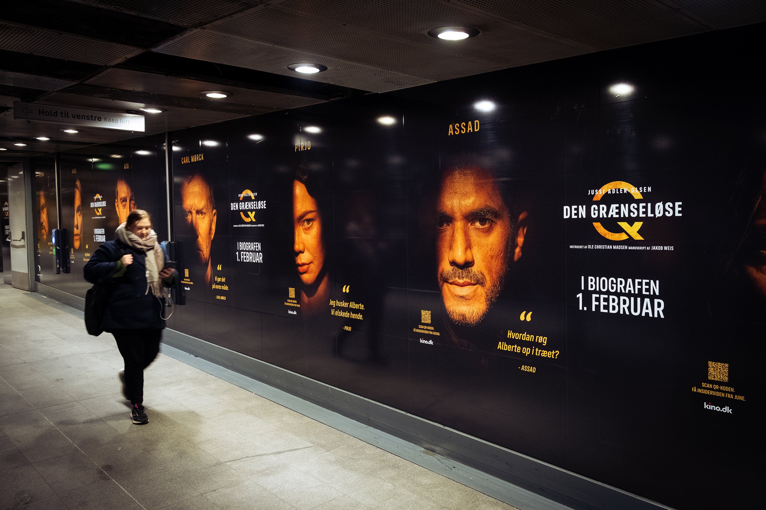 En person går gennem en metrostation, hvor der er store reklamer for filmen "Den Grænseløse" med portrætter af personer og tekst på dansk.