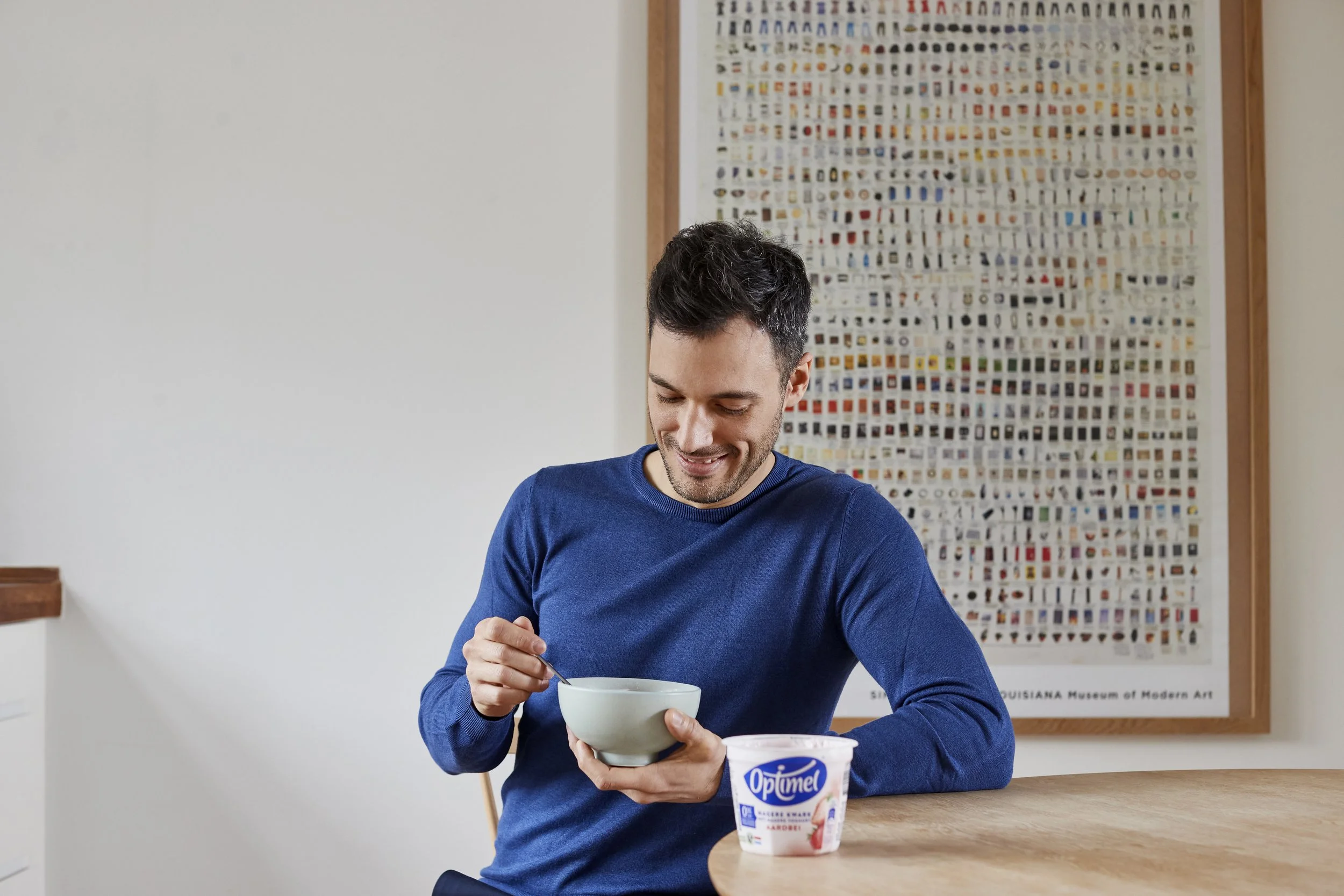 En mand i blå sweater sidder ved et træbord, smiler og spiser af en skål med optiMe yoghurt, der står foran ham.