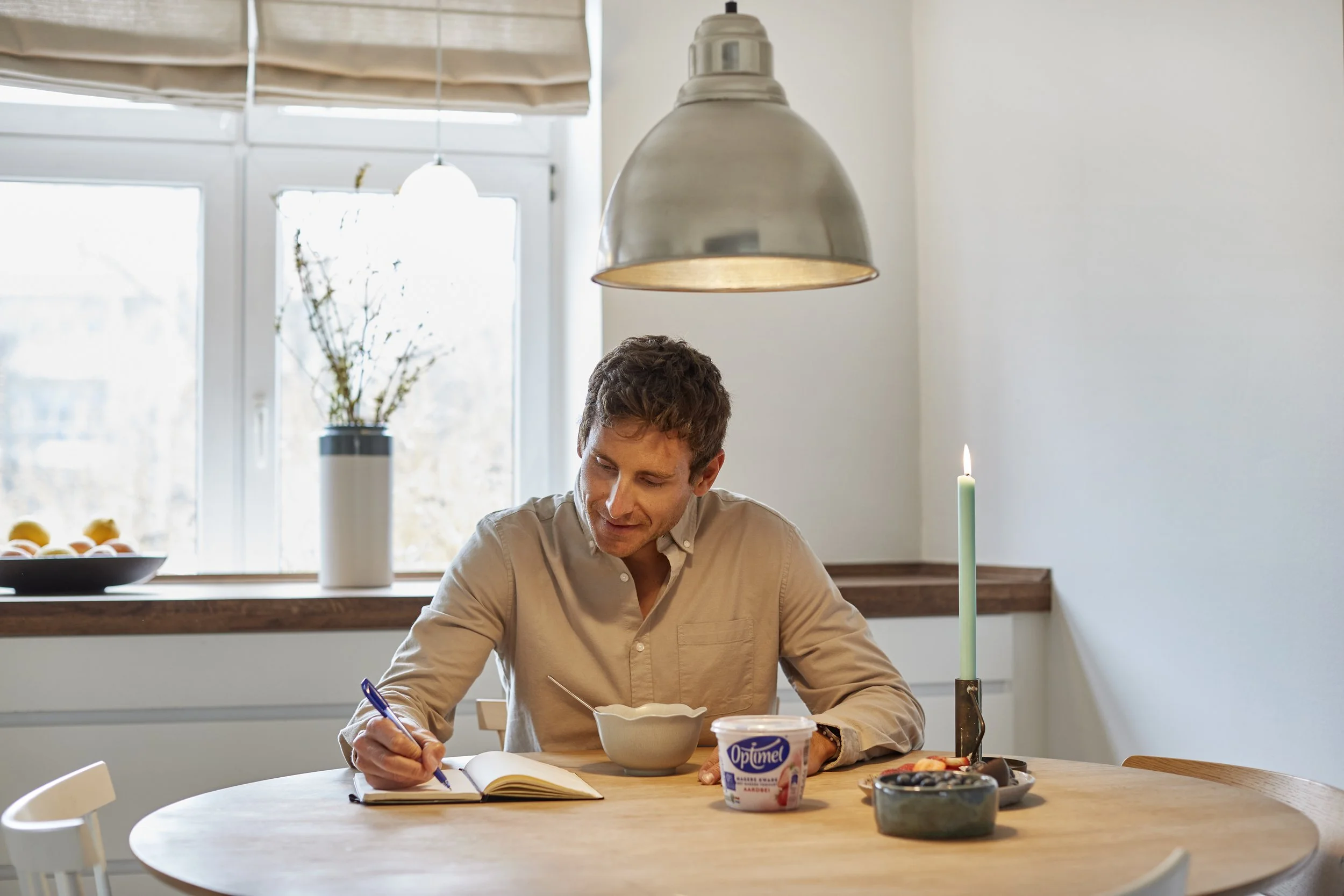 Mand sidder ved et rundt træbord, skriver i en notesbog, der er en skål med yoghurt, en lysestage med en tændt stearinlyst, en skål med blommer eller druer, og en vindueskarm i baggrunden. Der hænger en stor metalpendellampe over bordet.