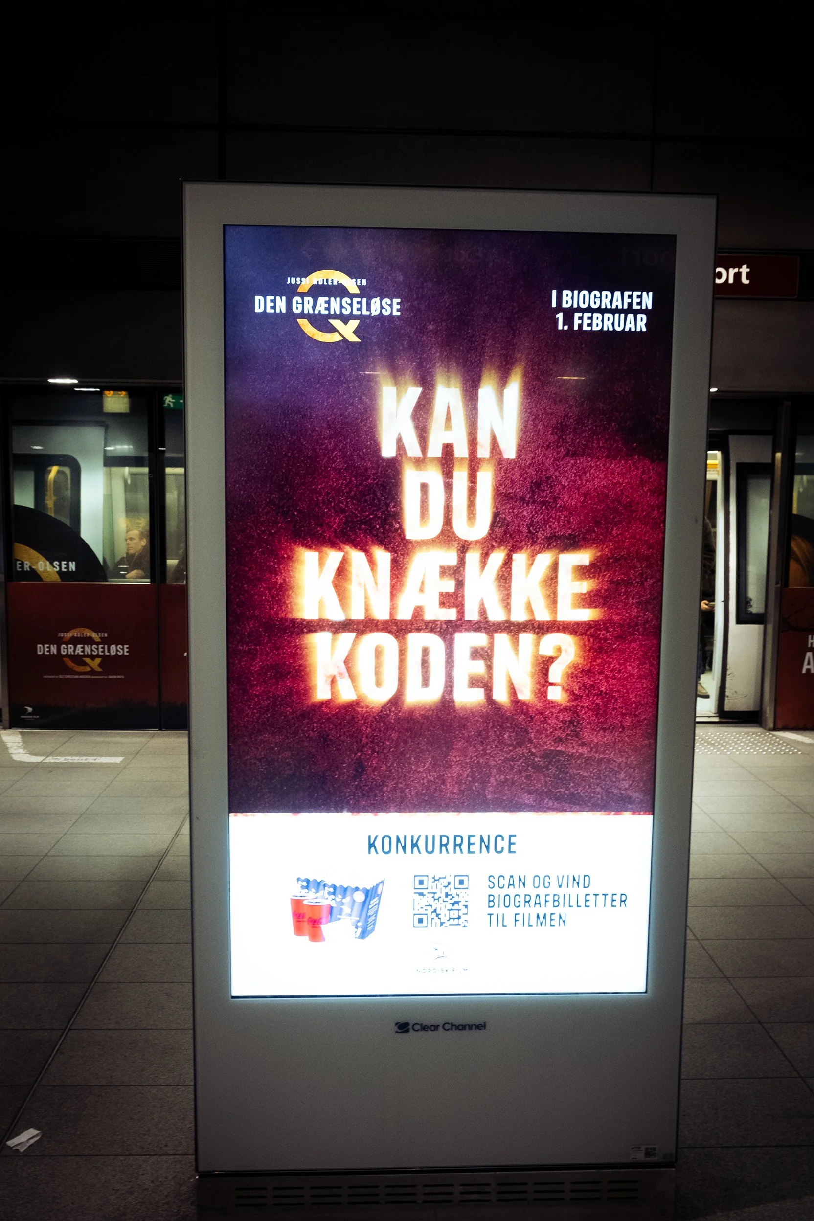 Reklameskilt i en togstation, der promoverer filmen 'Den Grænseløse' med teksten 'Kan du knække koden?'.