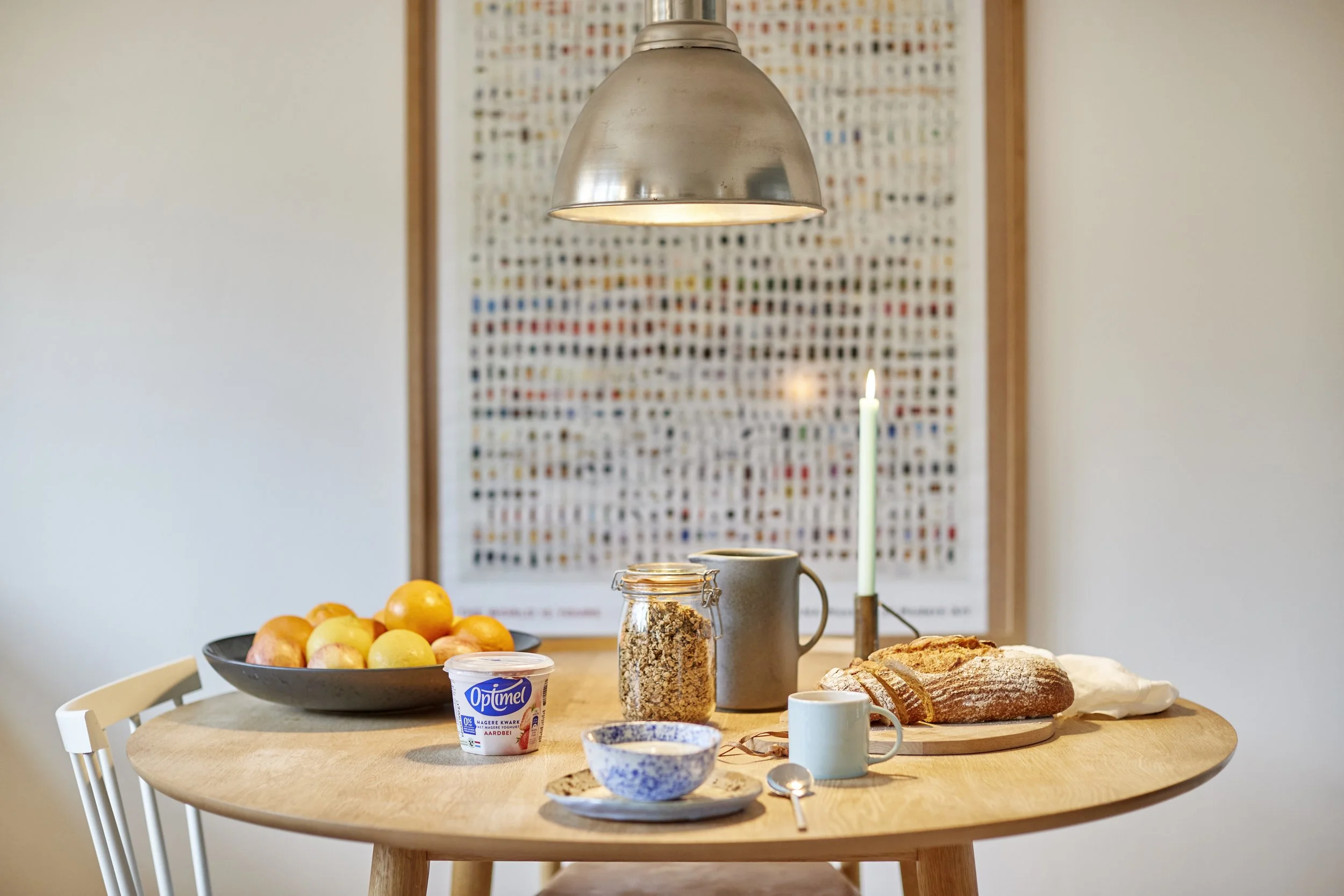 A round wooden dining table med frugt, yoghurt, kande med korn, kopper, skål med mad, og et brød. Der er også et lys i en holder og en tallerken med en ske. Bagved er der et vægkunstværk.