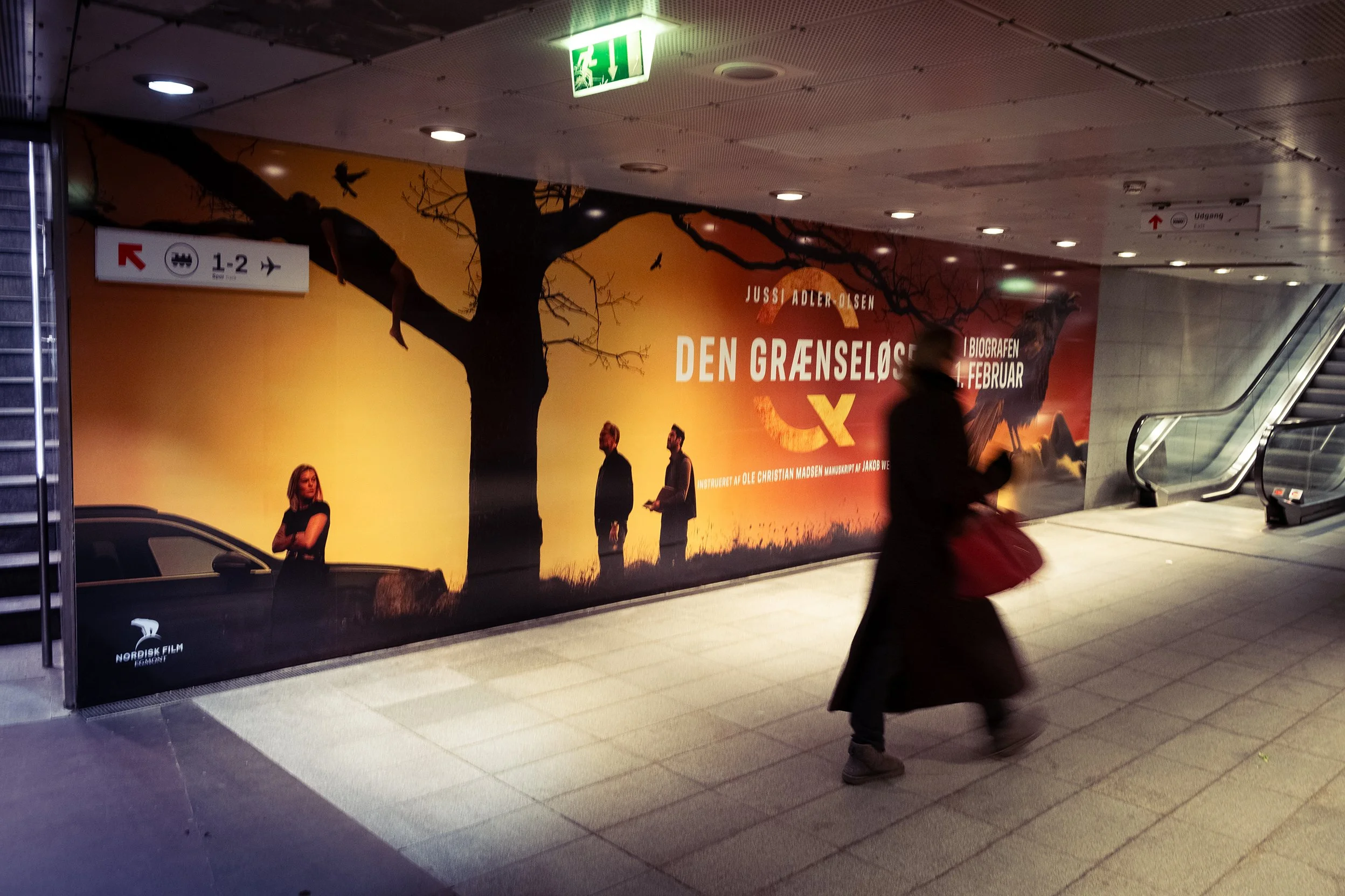 Person går forbi en reklamevæg i en underjordisk metrostation. Reklamen er for filmen 'Den Grænseløse' med Jussi Adler-Olsen og Oscar Wilde, og den er i orange og sort farver med en stor træsilhuet.