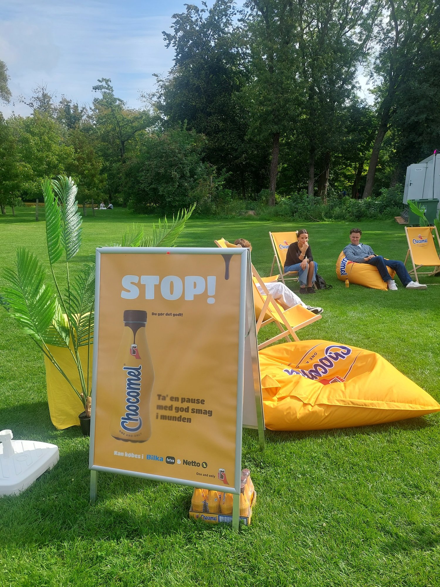 En reklameskilt med teksten STOP! og en flaske Chocomel, opstillet på en grøn parkeringsplads med grønne planter ved siden af. Der er liggende og siddende personer i baggrunden, samt store orange Chocomel-krus og solsenge.