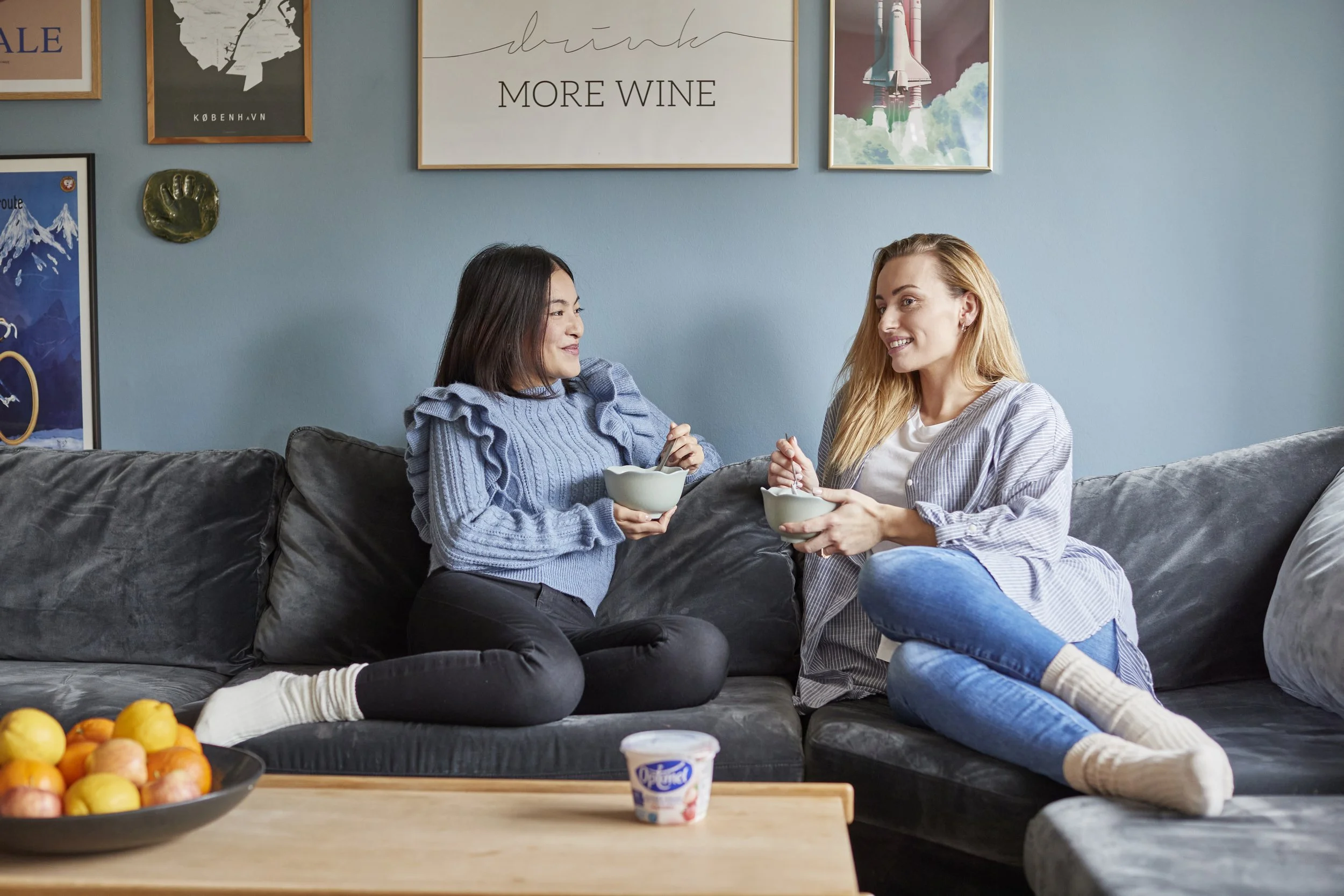 To unge kvinder sidder sammen på en sofa, spiser yoghurt og taler i et hjemligt rum med blå væg og billeder på væggen.