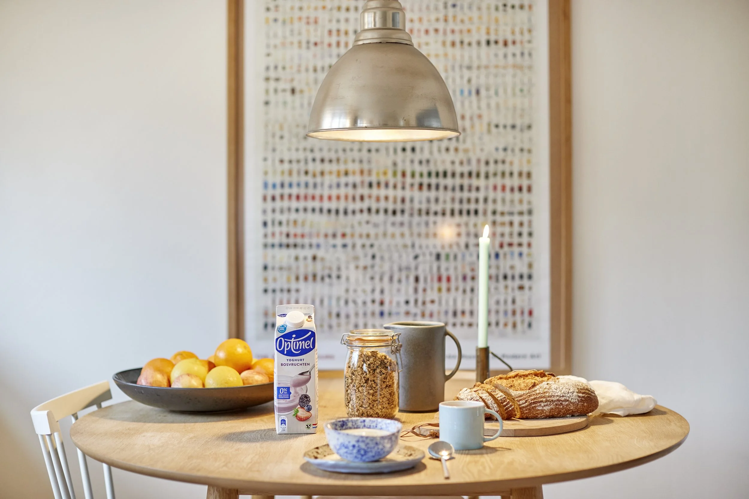 Morgenbord med frugt, yoghurt, mælk, granola, brød, kop, ske, lys, og tallerken på et rundt bord i et hyggeligt rum.