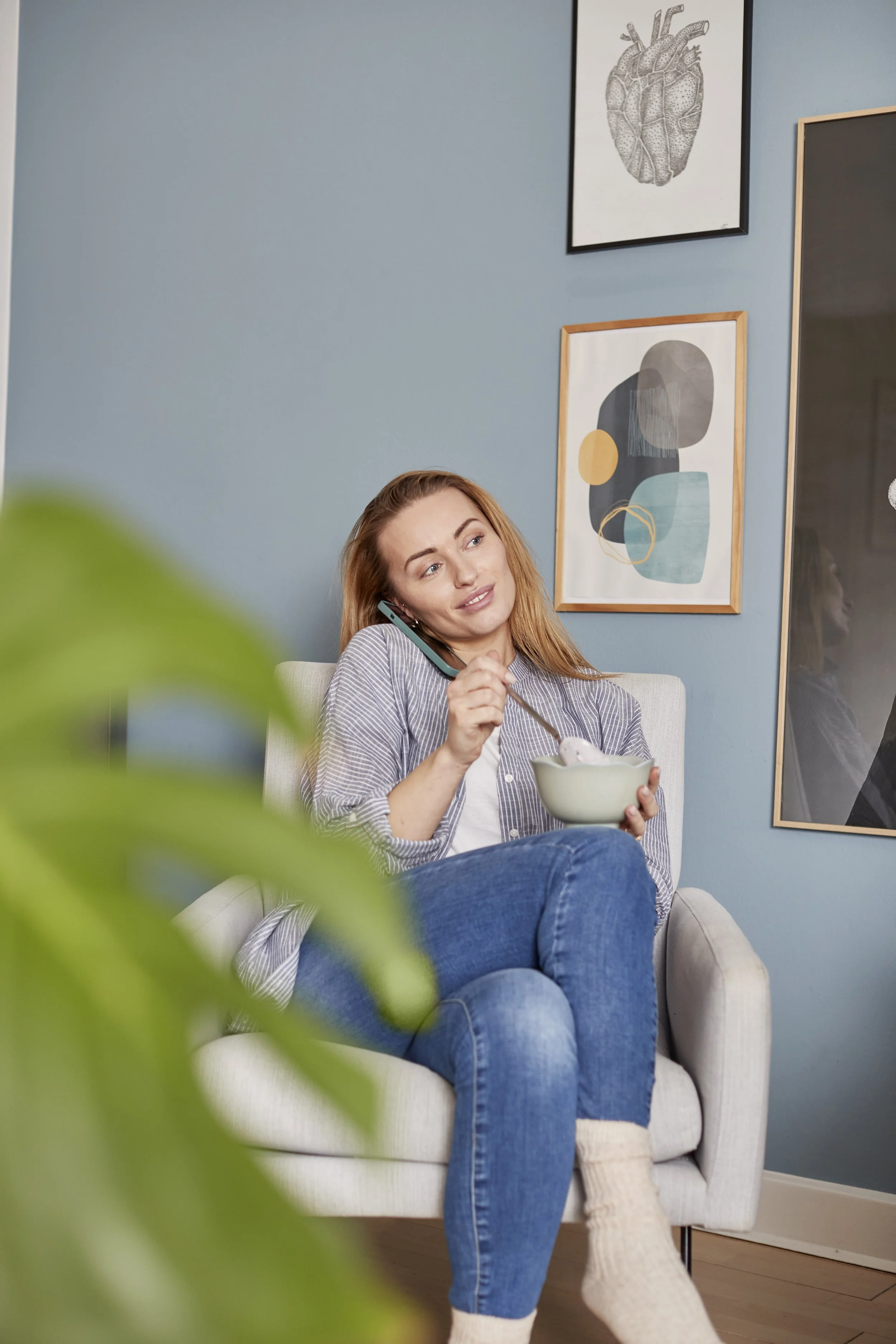 Kvinde sidder i en lænestol, spiser yoghurt, taler i telefon, og ser ud til at have en afslappende stund i sit hjem. Der er billeder på væggen bag hende og en grøn plante i forgrunden.
