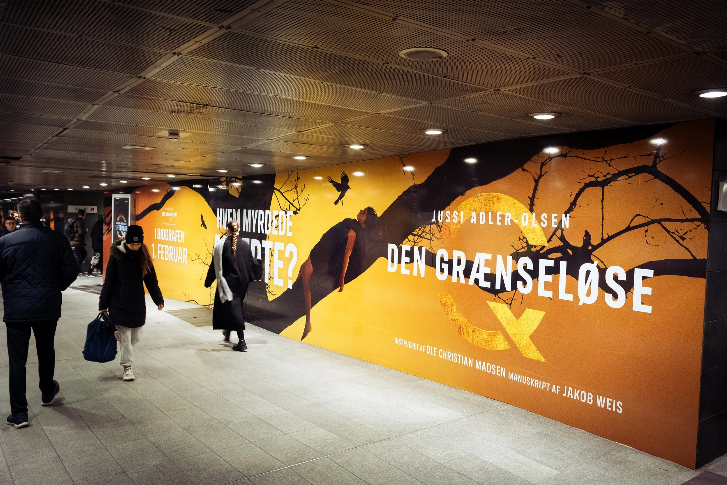 Annonce i metrostation med orange baggrund, grafik af en gren og fugle, tekst om bogen 'Den Grænseløse' af Ussi Adler-Olsen, med information om forfatter og udgivelse.