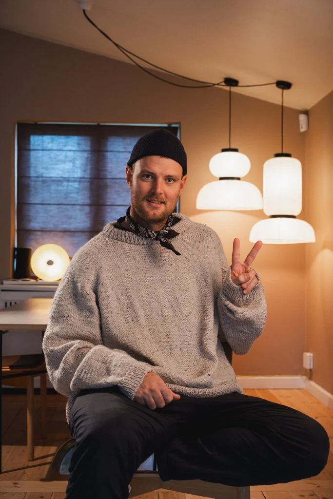 Ung mand sidder på en stol i et hjem, iført en grå sweater, sort hue og laver en peace-tegn med hånden. Bag ham er moderne lyspendler og et vindue dækket med mørke persienner.