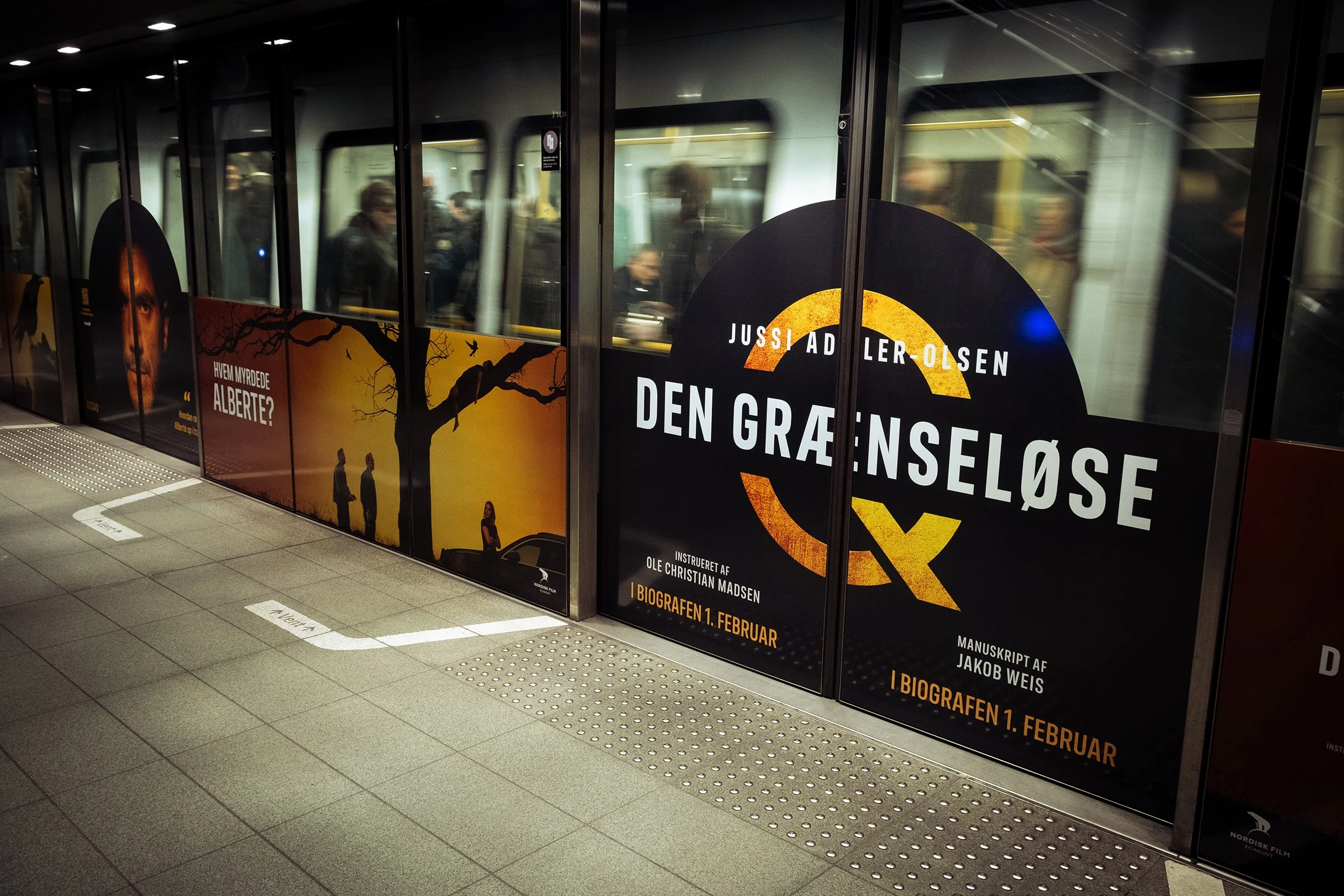 Advertisement for the film 'Den Grænseløse' in a metro station, with a large poster displaying the title, director's name, and screening date. The poster features an illustration of a tree and silhouettes of people.
