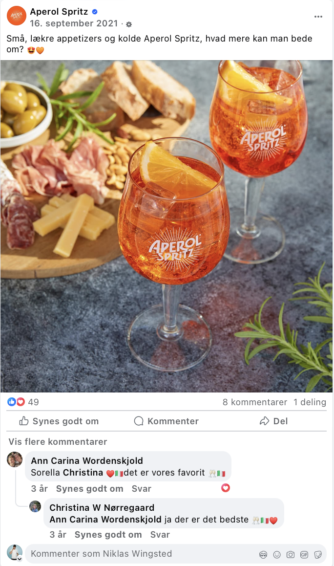 To glas Aperol Spritz med skiver af citron, foran en tapasbræt med ost, skinke, oliven, brød, og pølser.