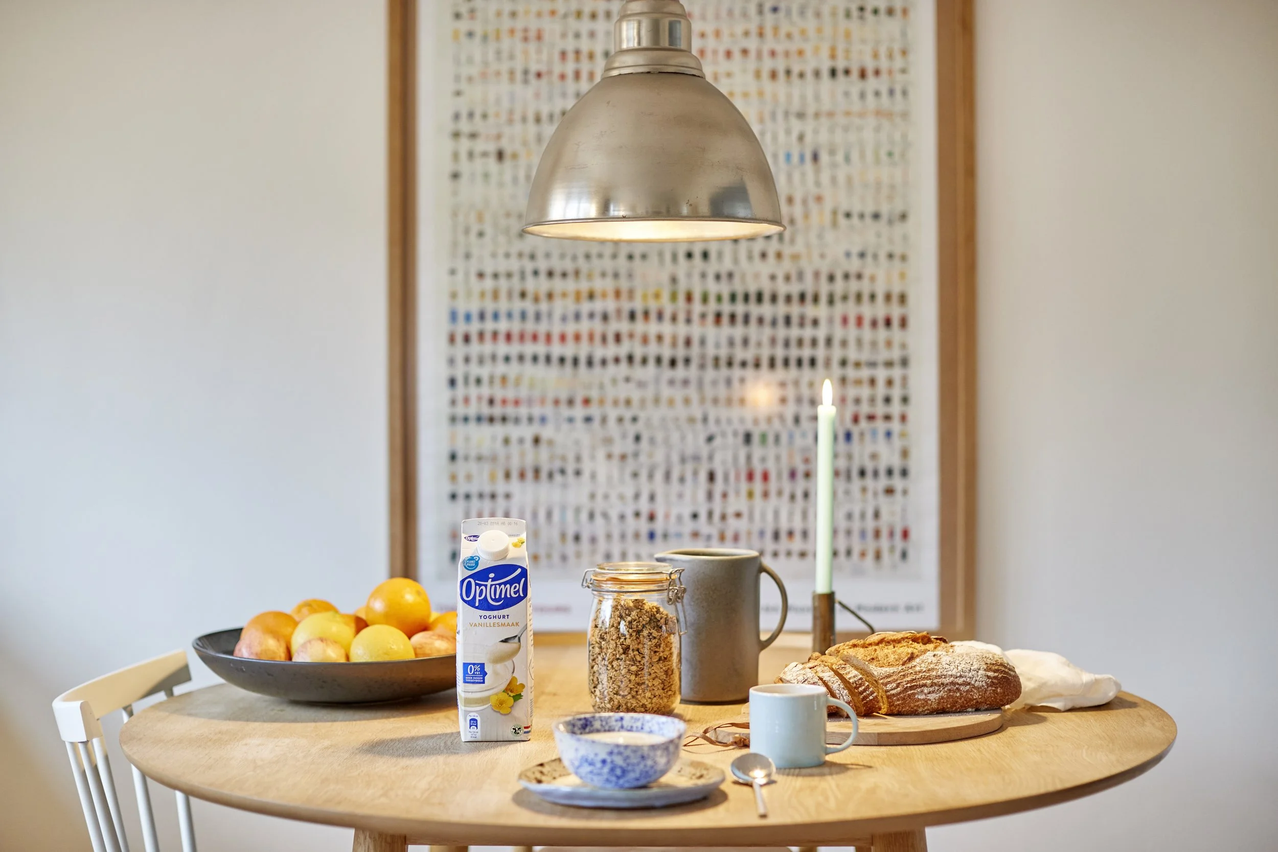 Morgenbord med frugt, yoghurt, brød, kop, ske, tændt stearinlys på træbord.