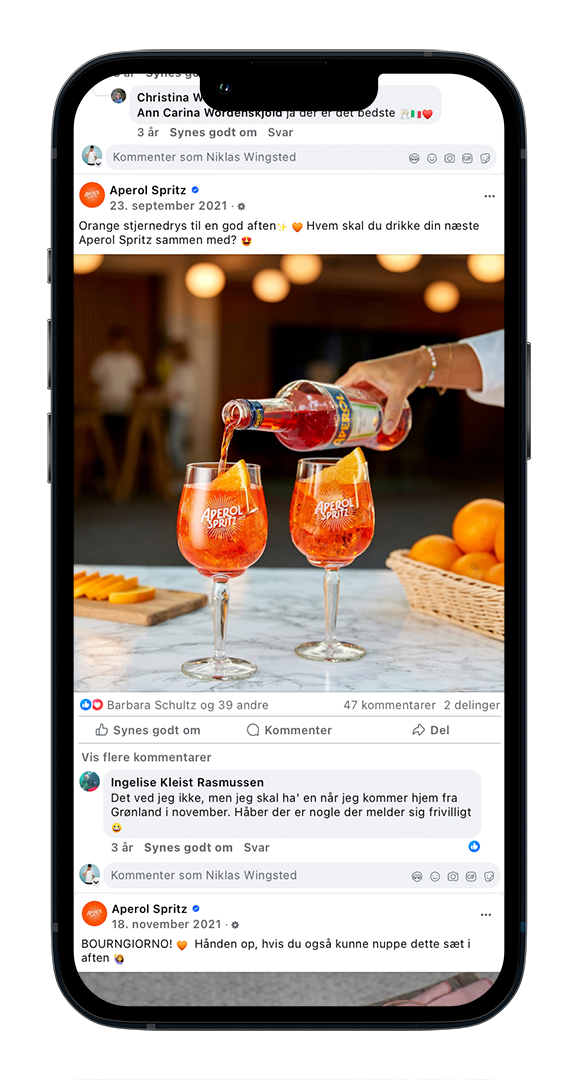 Et glas Aperol Spritz bliver fyldt med aperol likør på en barbord, omgivet af appelsiner og baggrund med bløde lys. En person holder flasken og hælder likøren i glasset.