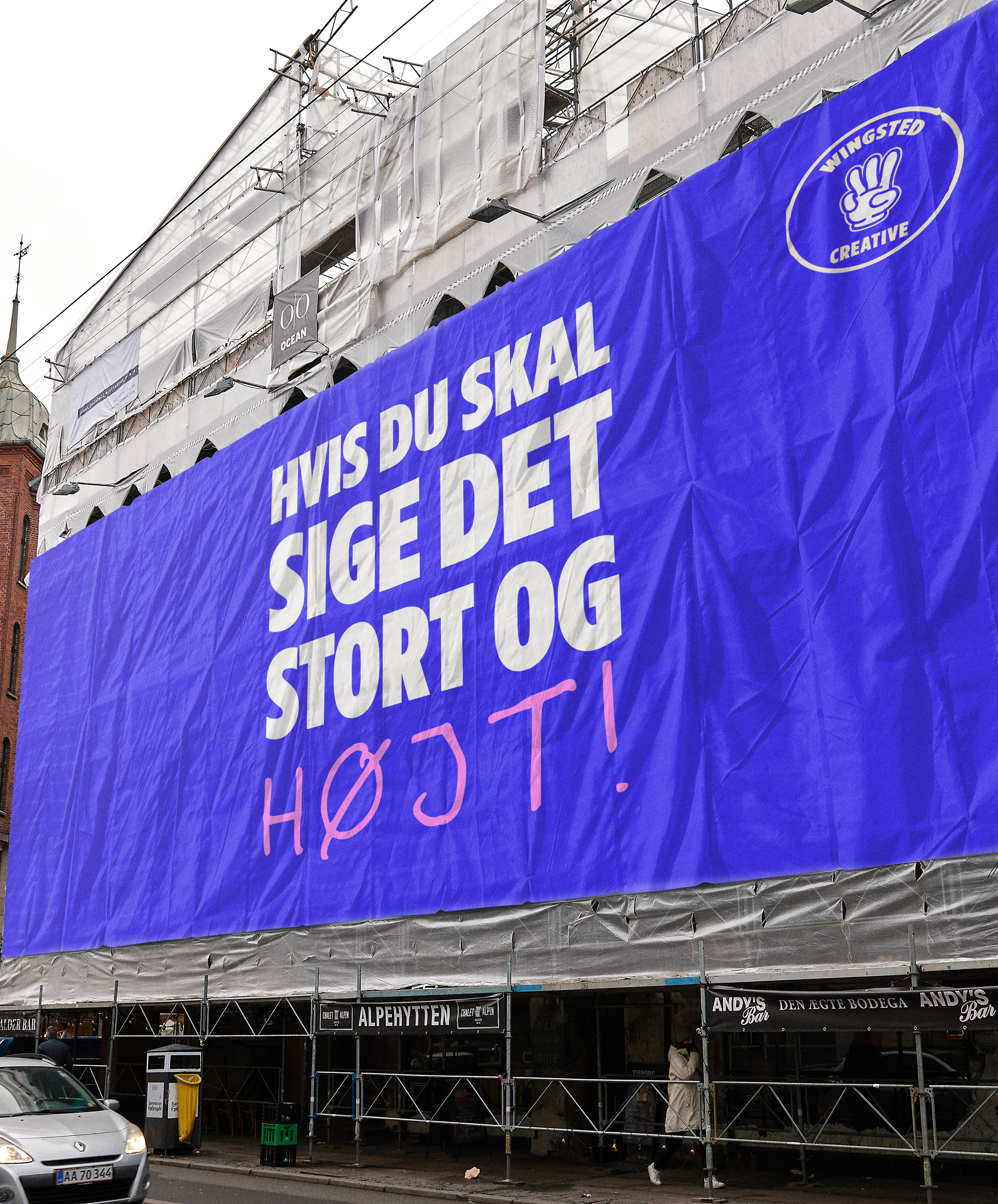 Stor blå banner med hvid tekst, der siger: "Hvis du skal sige det stort og højt!" på en bygning under renovering, og et logo med en hånd, der laver en v' håndtegn, ved navn "Wingsted Creative."