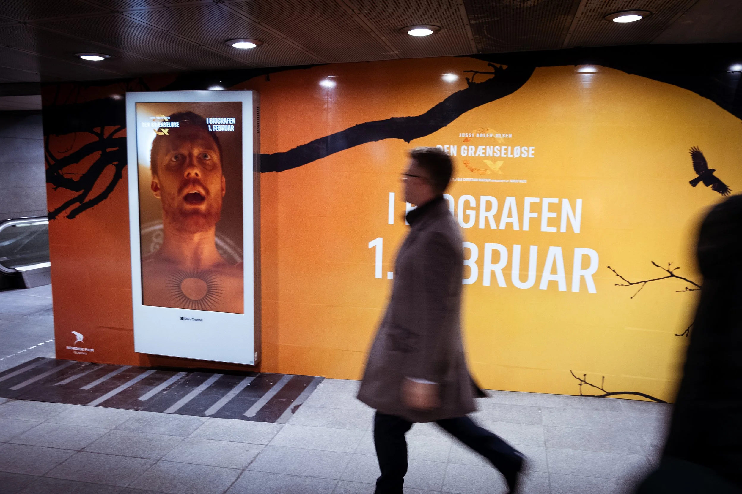 En reklameplakat for filmen 'Den Grænseløse' med en mand, der har åbent mund og synlige halsår, placeret i en metrostation. Baggrunden er orange med tekst om filmens premieredato.