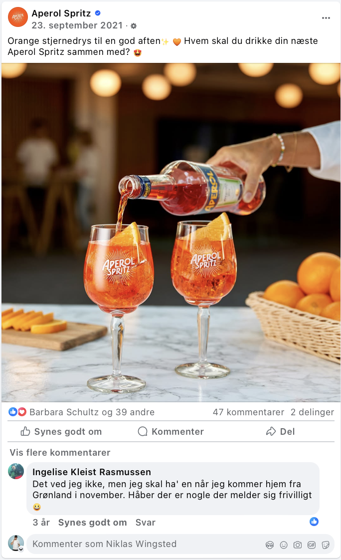 To glas med Aperol Spritz, der indeholder is og appelsinskiver, en person hælder Aperol fra en flaske i glassene, der står på en bord med appelsiner og ostetilbehør.