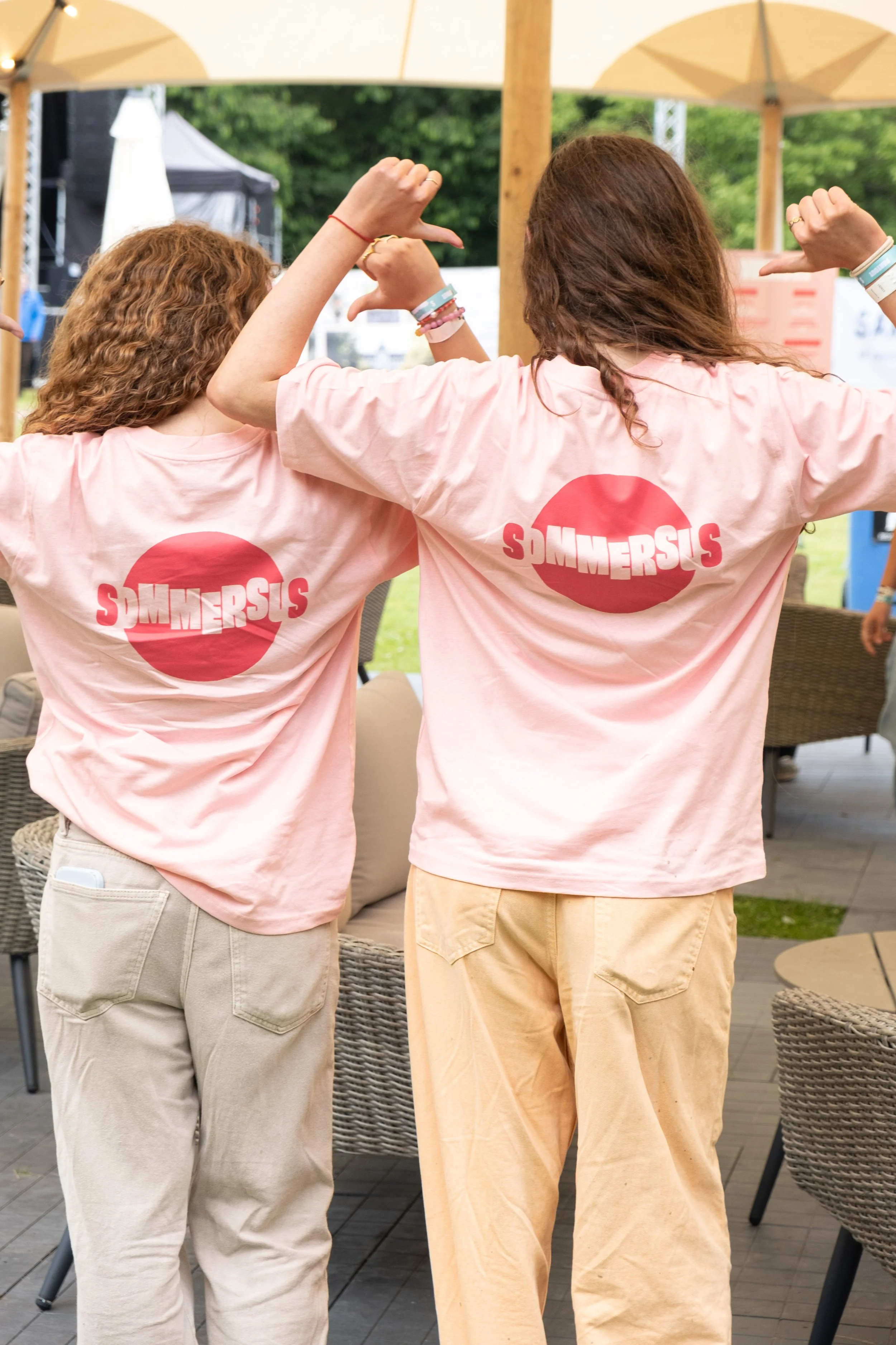 To personer med lyserøde T-shirts, hvor der står 'Sommersis', står med ryggen til og laver mavebøjning udendørs under pergola.