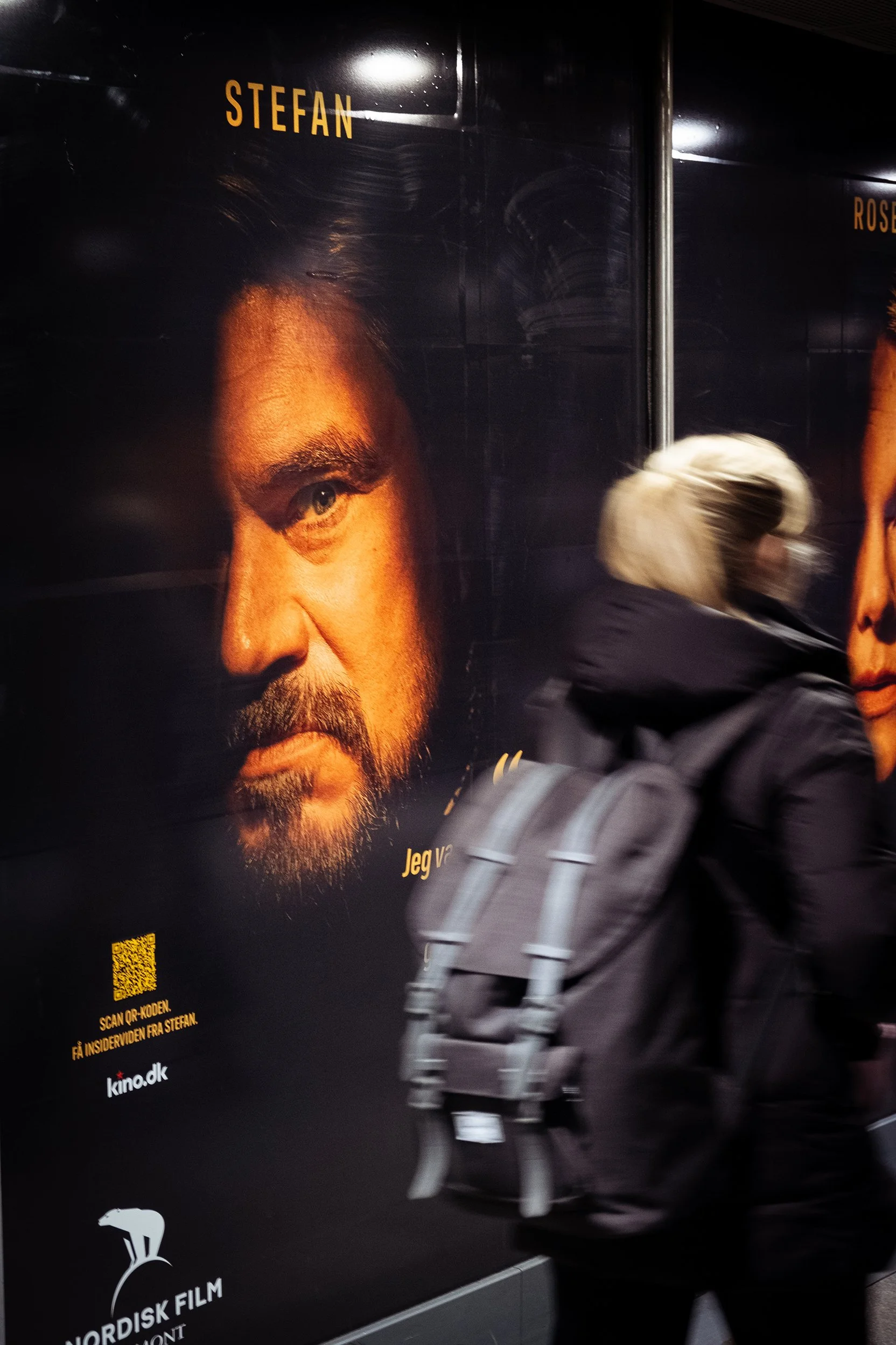 En person med blondt hår og backpack går forbi en reklameplakat med portræt af en mand med skæg og mørkt hår. Tekst på plakaten inkluderer navnene Stefan og Ros.