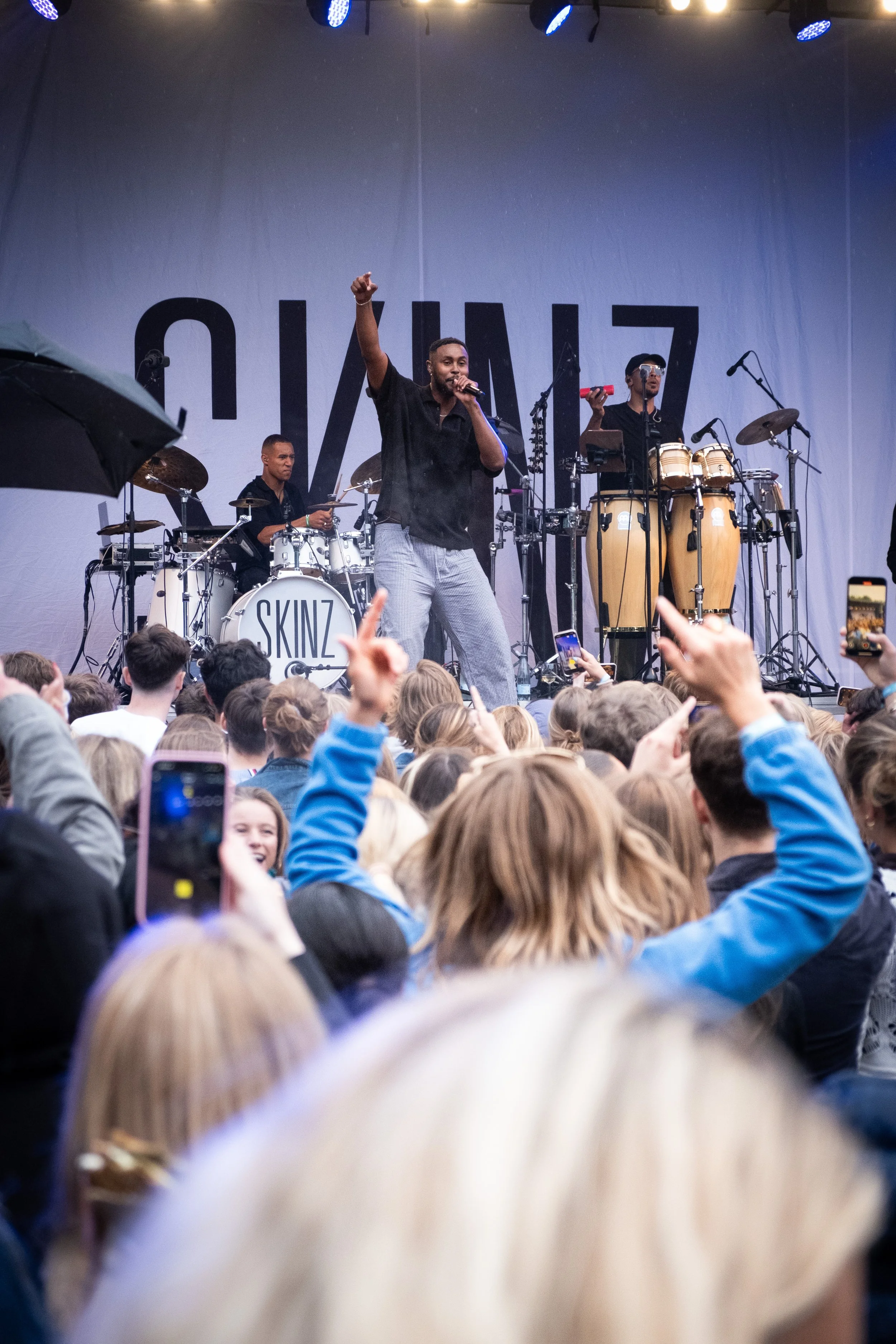 En musikgruppe optræder på scene foran et publikum, med en sanger i midten, en trommeslager og percussionmusikere, der spiller sammen, med en stor gruppe mennesker, der ser på og tager billeder.