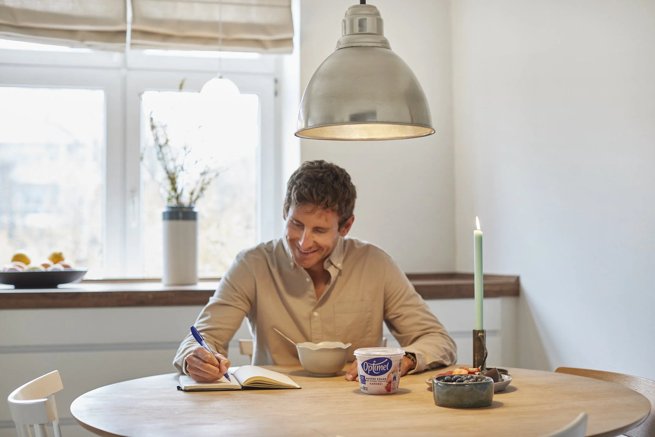 En mand sidder ved et rundt spisebord, skriver i en notesbog med en blå pen, med en skål ylette yogurt og en tallerken med bær på bordet, mens han smiler. Der er en tallerken med æg på vindueskarmen i baggrunden.