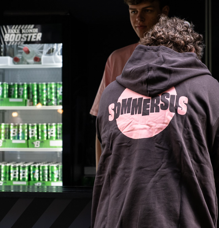 To personer står foran en vending maskine med grønne sodavand. En person har en hættetrøje med teksten 'SUMMERSUS' på ryggen.