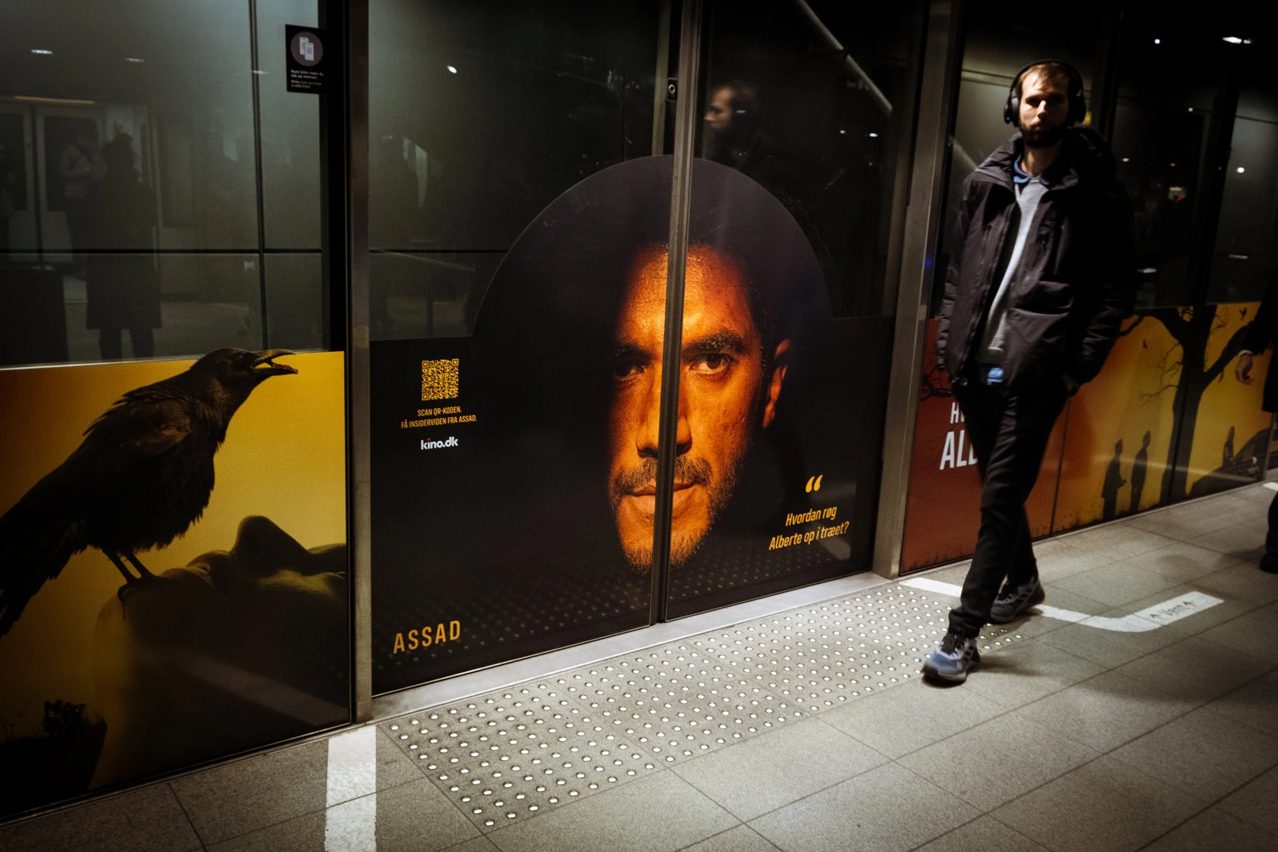 En person står med krydsede arme ved en metroindgang med reklamer. En stor reklame viser en mand med mørk hud og kort hår. Der er også en reklame med en sort fugl på venstre side.