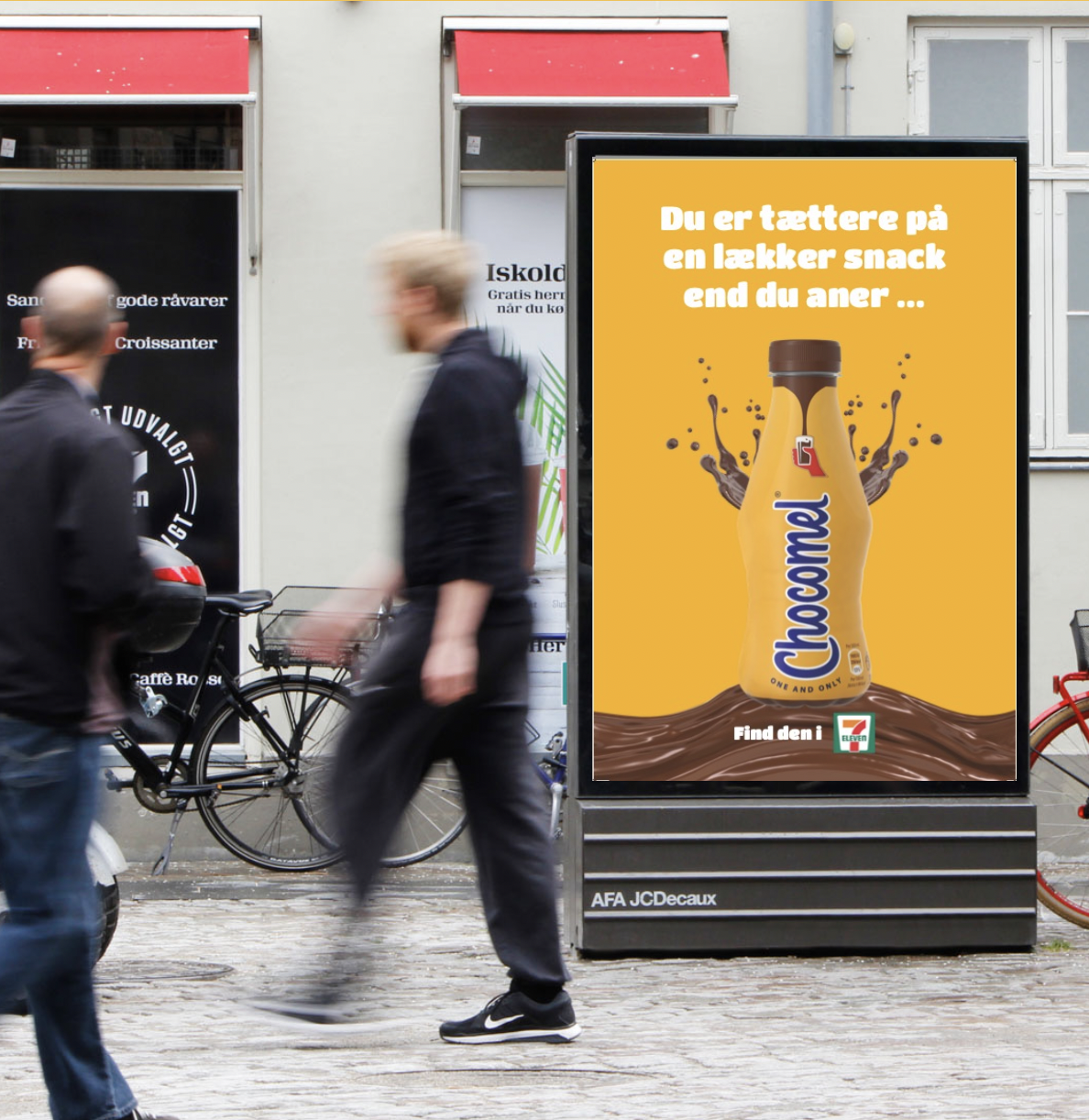 En reklamesøjle for Chocomel med teksten, 'Du er tættere på en lækker snack end du aner ...' og er grafisk udformet med en chokoladesmurret flaske på en gul baggrund.