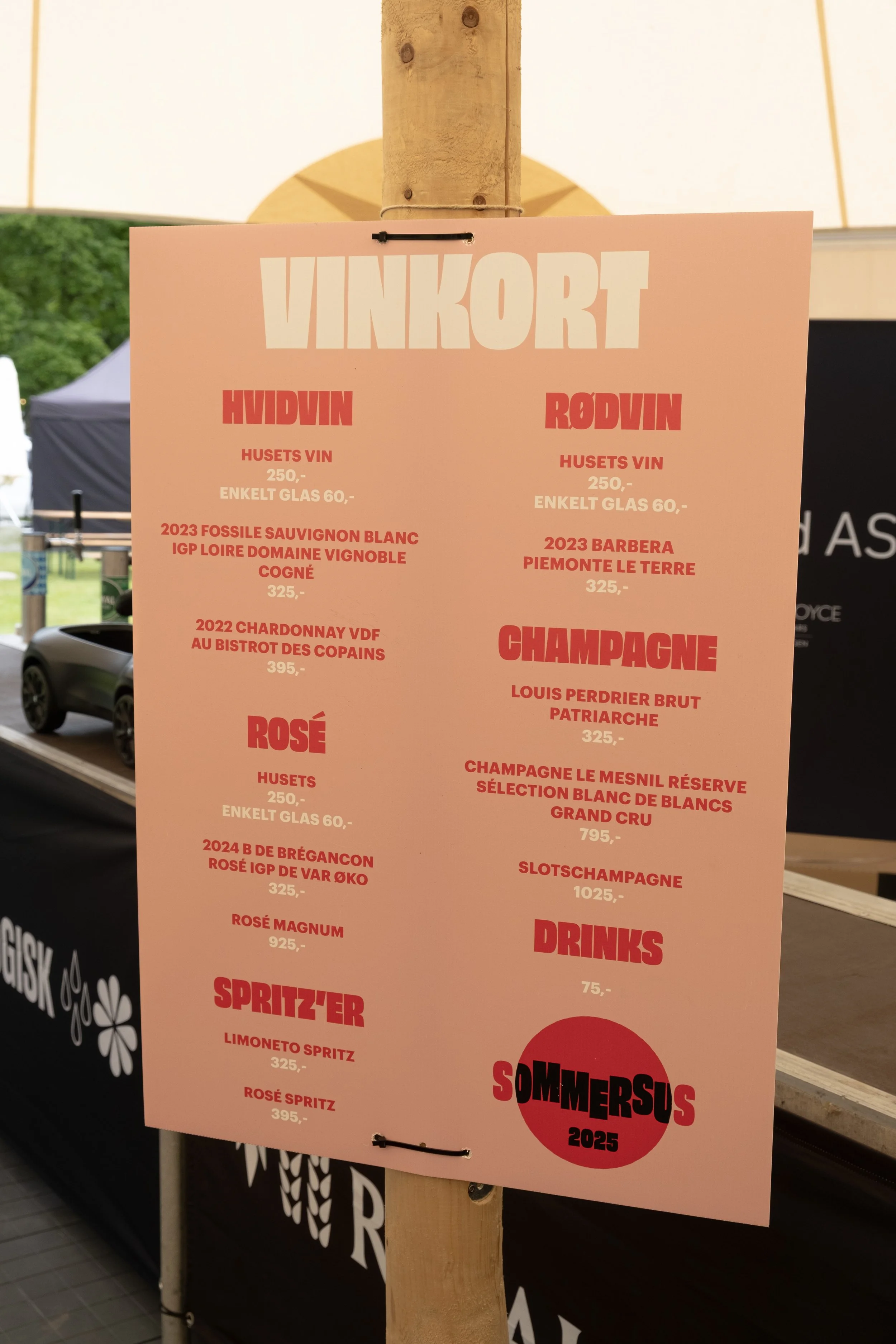 En menuplakat med vin, champagne, rosé, drinks og priser, inddelt i kategorier for Hvidvin, Rødvin, Champagne, Rosé og Drinks. Baggrund med grønne træer og en sort telt.