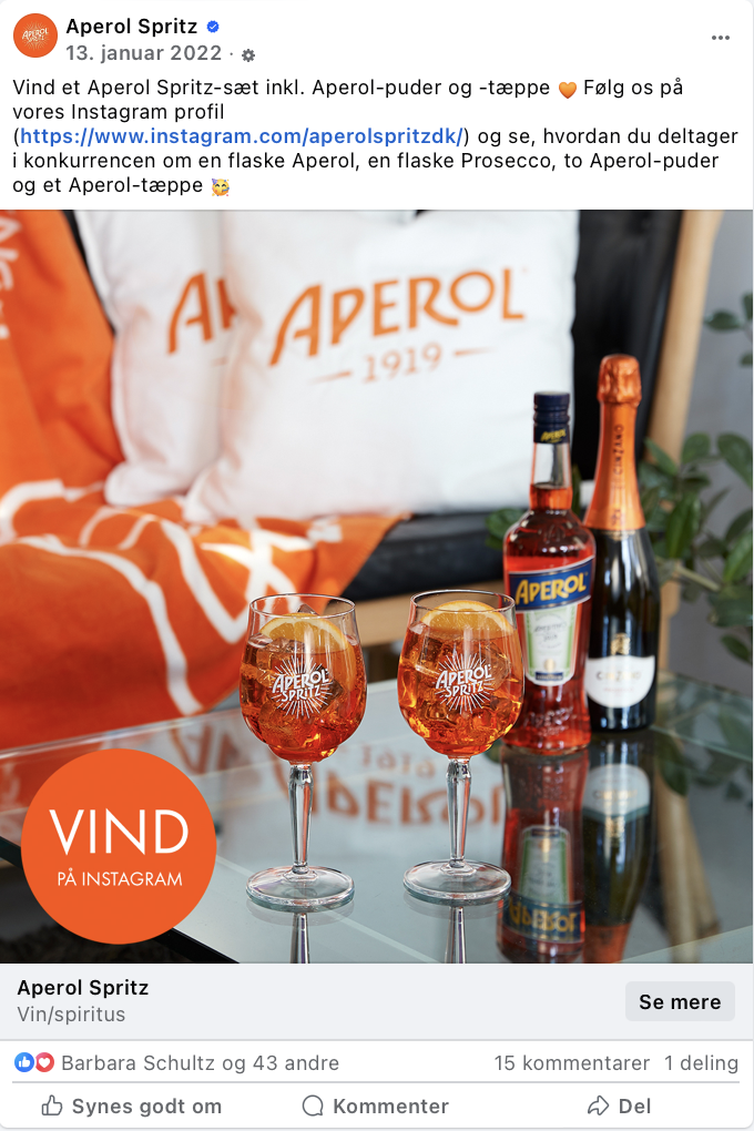 To Aperol Spritz-drikke i glas med Aperol-logo, to Aperol-flasker bagved, en stor Aperol-pude og et Aperol-tæppe i baggrunden.