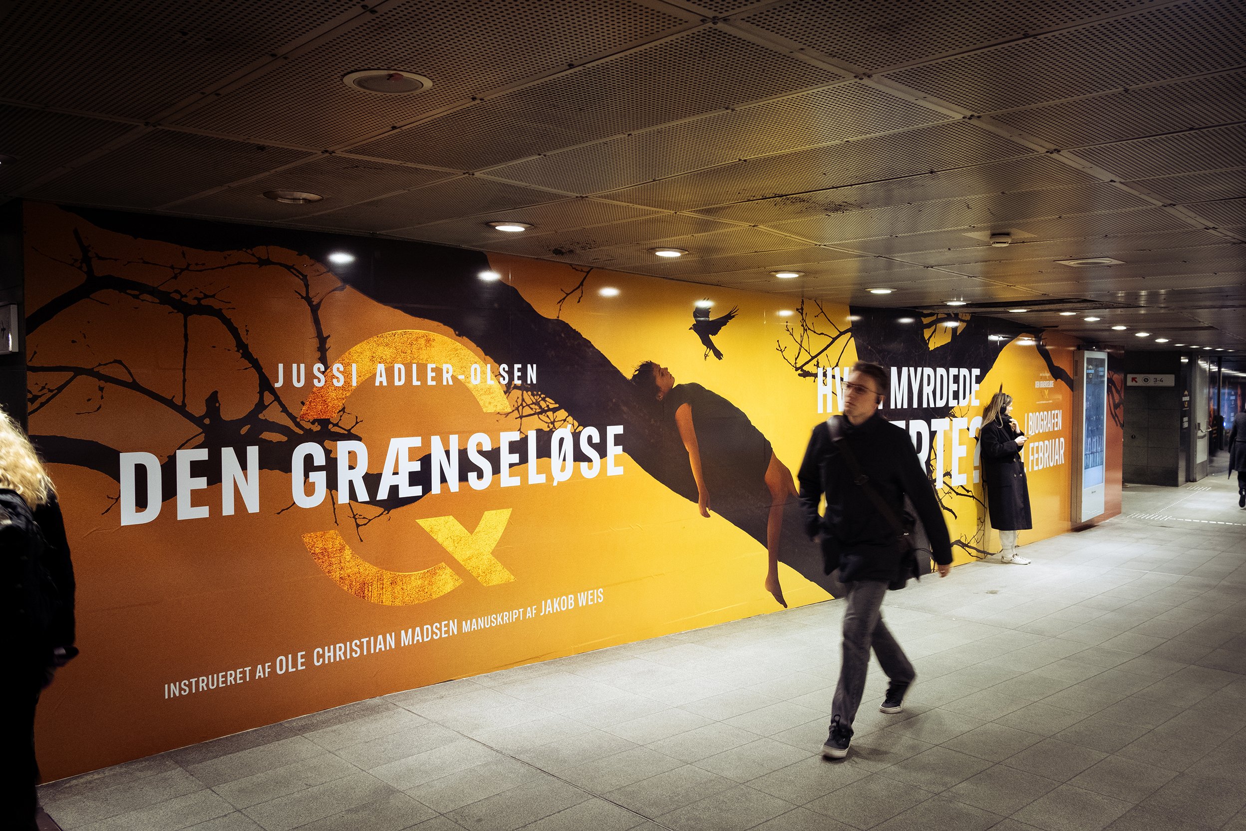 Kunstnerisk reklamevæg med sort trægren, en person hængende fra en gren, en flyvende fugl, et stort orange solopgang eller solnedgang, og tekst på dansk.