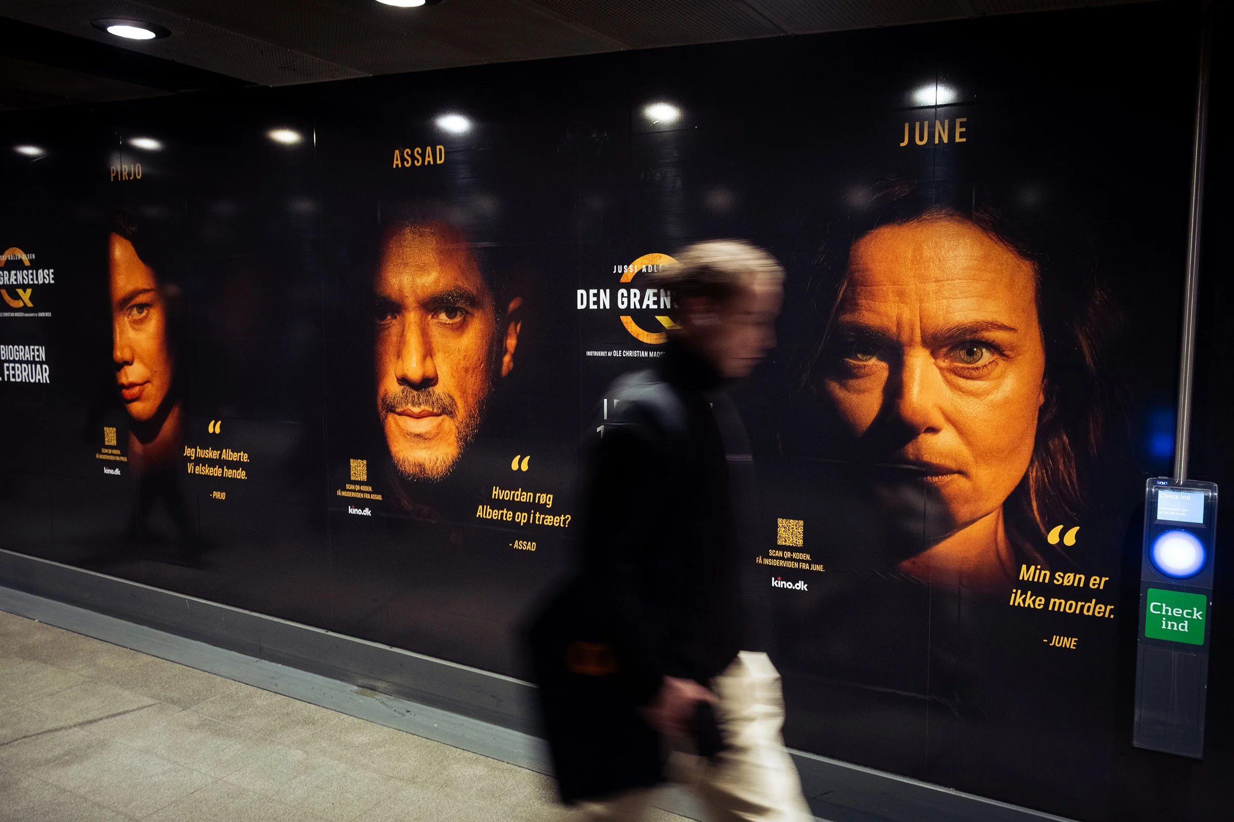 Person går forbi en reklamevæg i en metrostation med plakater for filmen 'Den grøn' med portrætter af skuespillere og citater oversat til dansk.