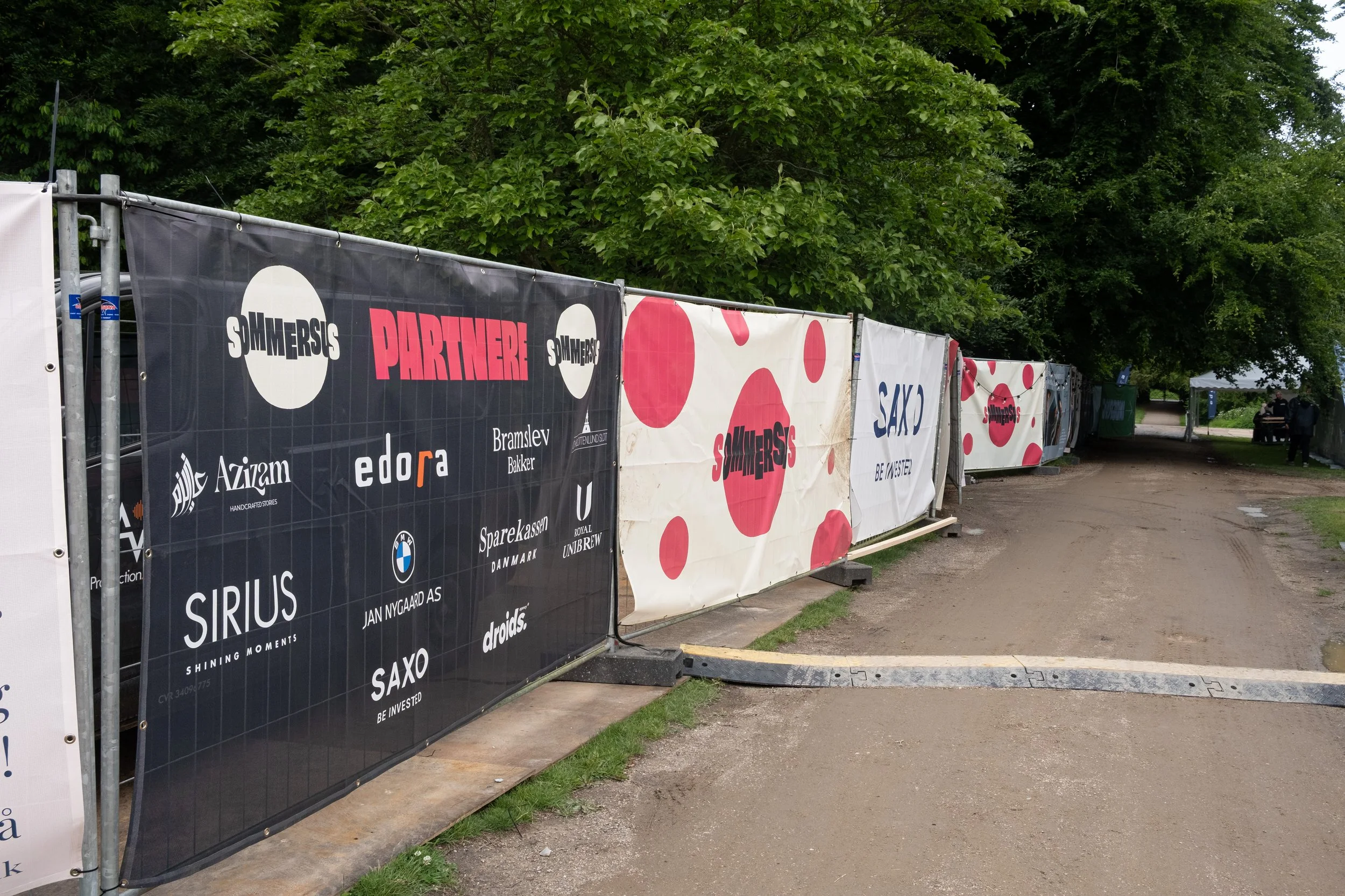 En række af reklameskilte på en støttemur ved en sti med træer i baggrunden.