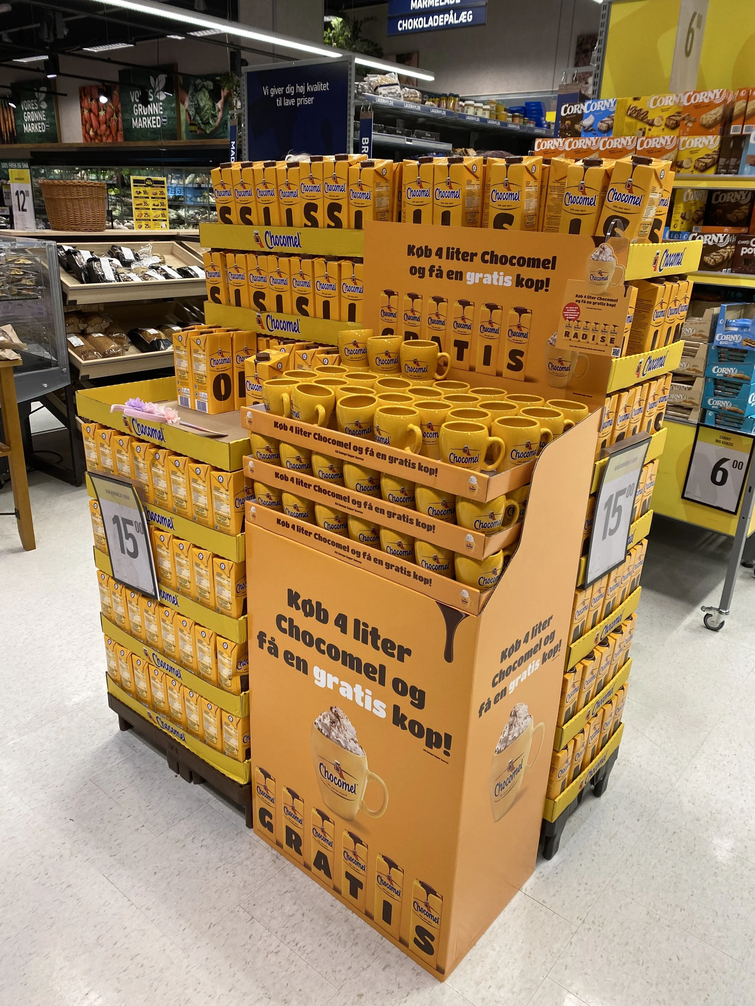 Salgsdisplay med Chocomel drikkevarer, køb 4 liter og få en gratis kop, tydeligt skilt. Opbevaringshylde med andre fødevarer i baggrunden.