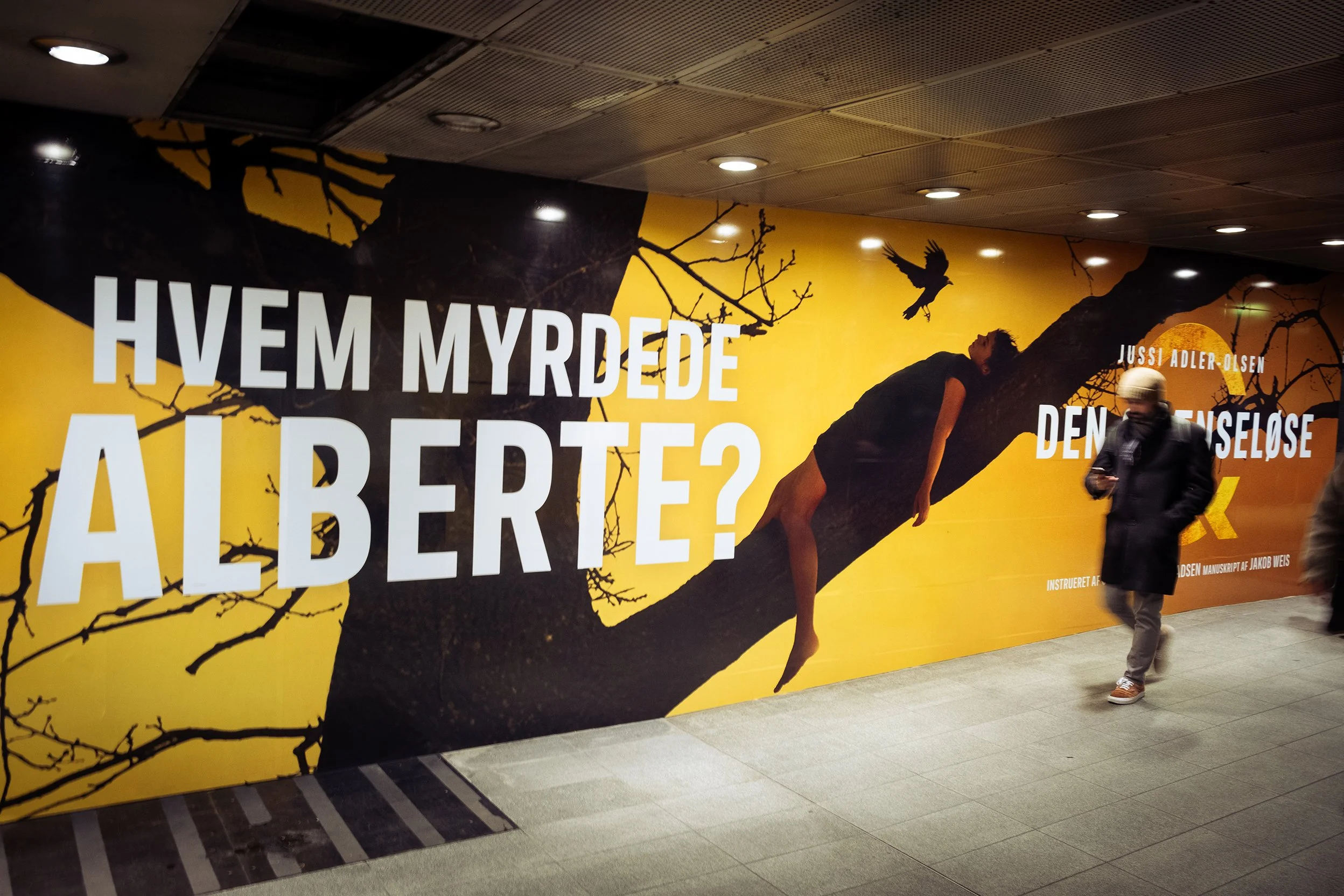 Grafisk væg med stor tekst: 'Hvem myrdede Al Berto?', og et kunstnerisk billede af en person hængende i en stor gren af et træ med en sort fugl i baggrunden, hvid tekst med forfattere og instruktion.