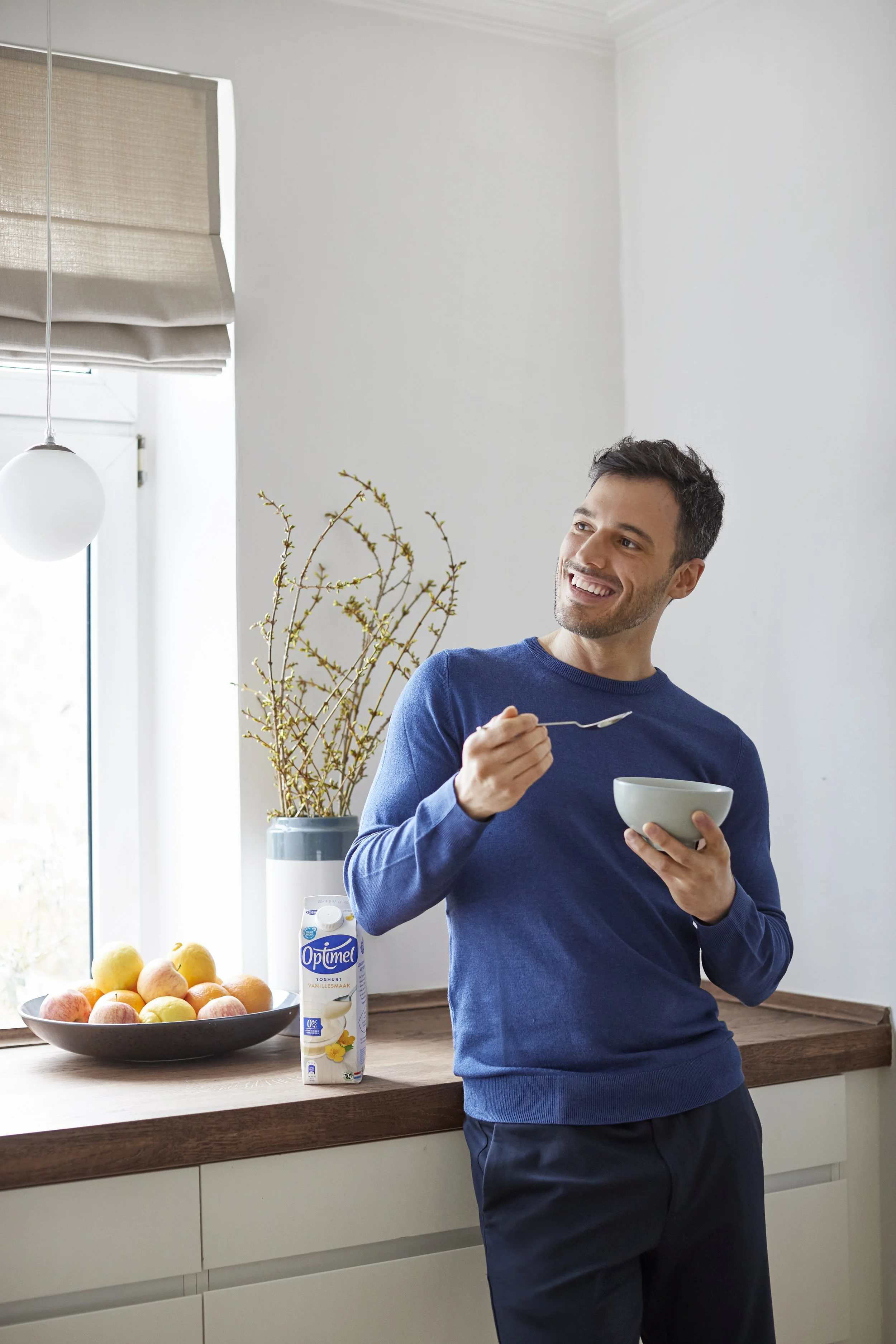 En mand i en blå sweater står i et køkken, smiler og spiser yoghurt med en ske. På bordet står en skål med frugt og en pakke yoghurt.