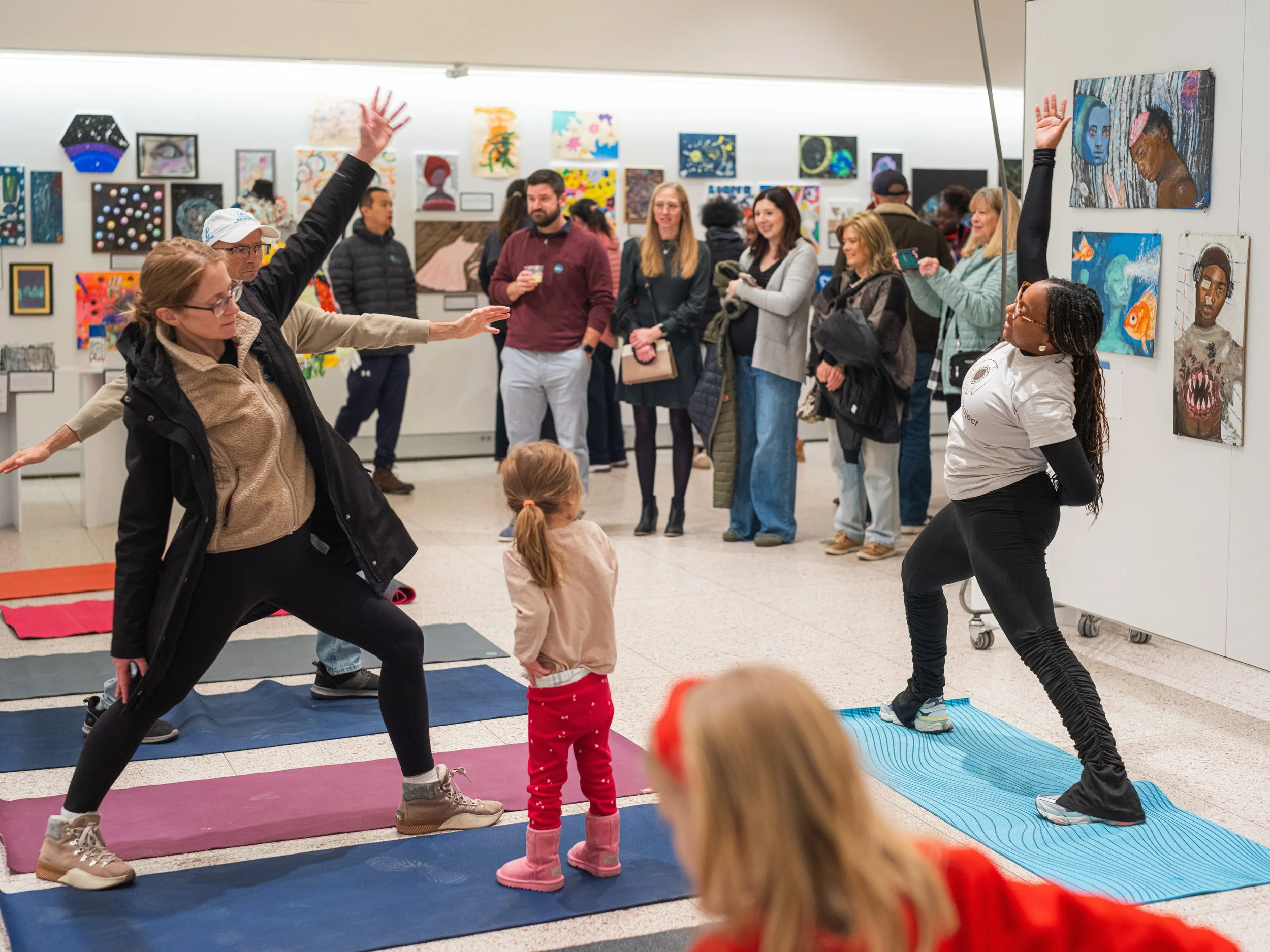 Two women leading a yoga class on mats with children, surrounded by art gallery visitors observing paintings.