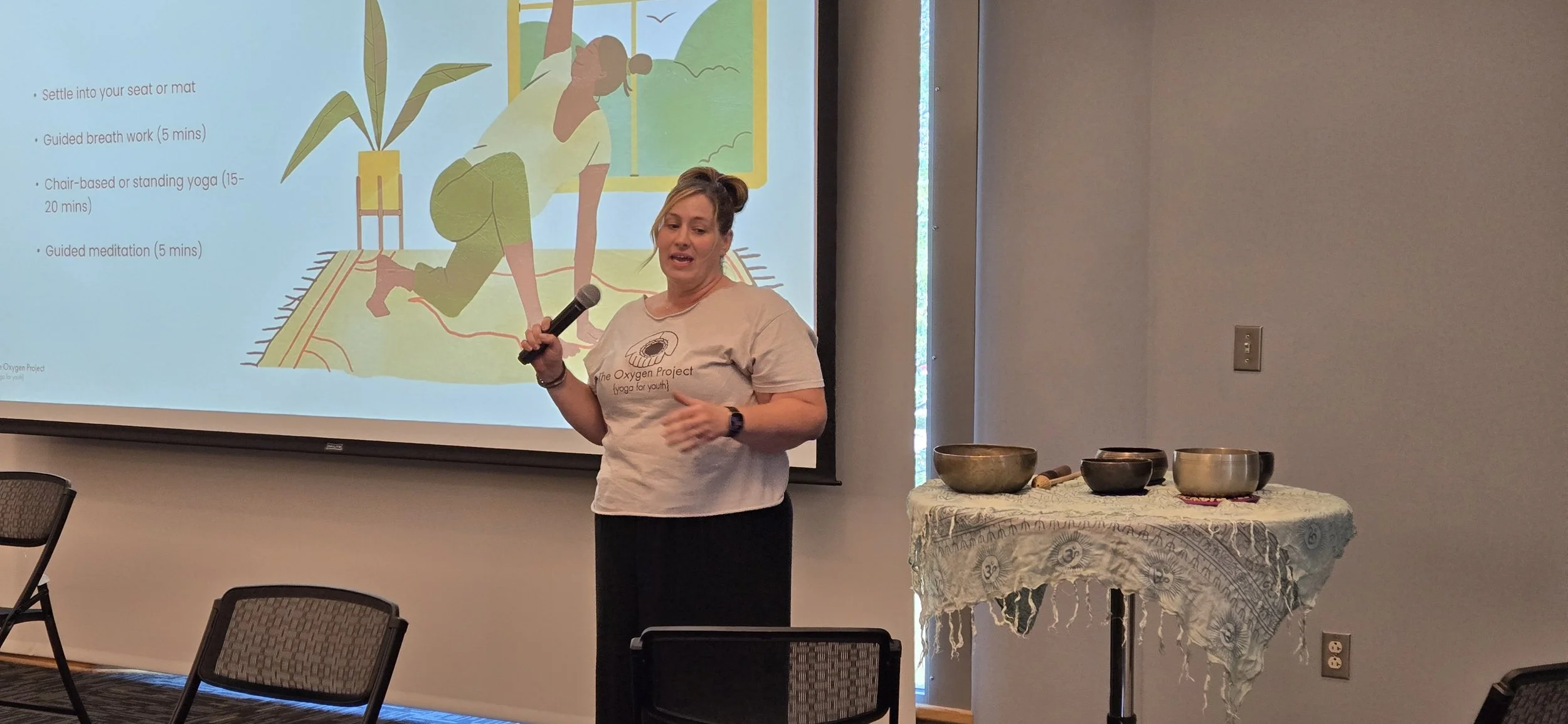 A woman giving a presentation about yoga and breathing exercises in a room with a large screen displaying illustrations of a person practicing yoga. She is holding a microphone and standing next to a table with singing bowls.
