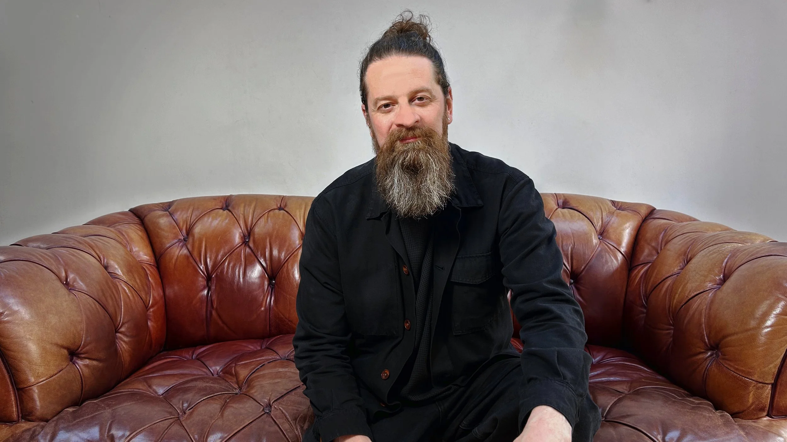 A man with a beard and hair tied up in a bun sitting on a vintage brown leather tufted sofa against a plain light gray wall.