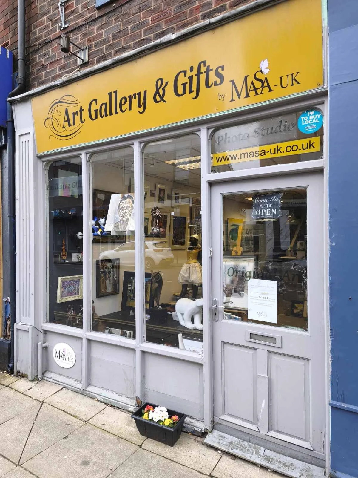 storefront with yellow sign reading "Art Gallery & Gifts by MASA-UK"; display window with art pieces and sculptures; grey door with "Come In We Are OPEN" sign; small flower pot outside.