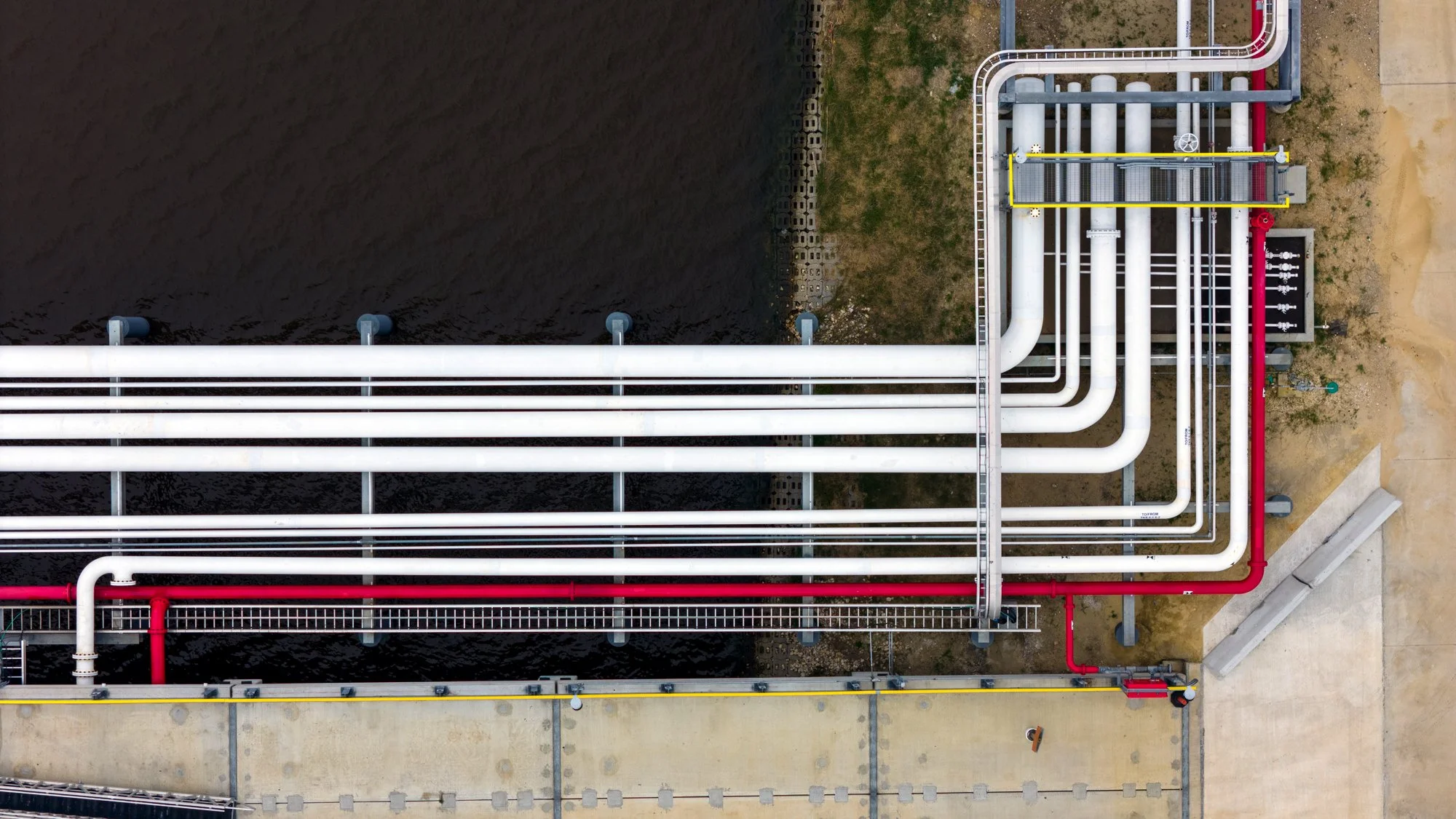 Aerial view of an industrial pipeline system with white and red pipes, valves, and metal supports over a water body and concrete ground.