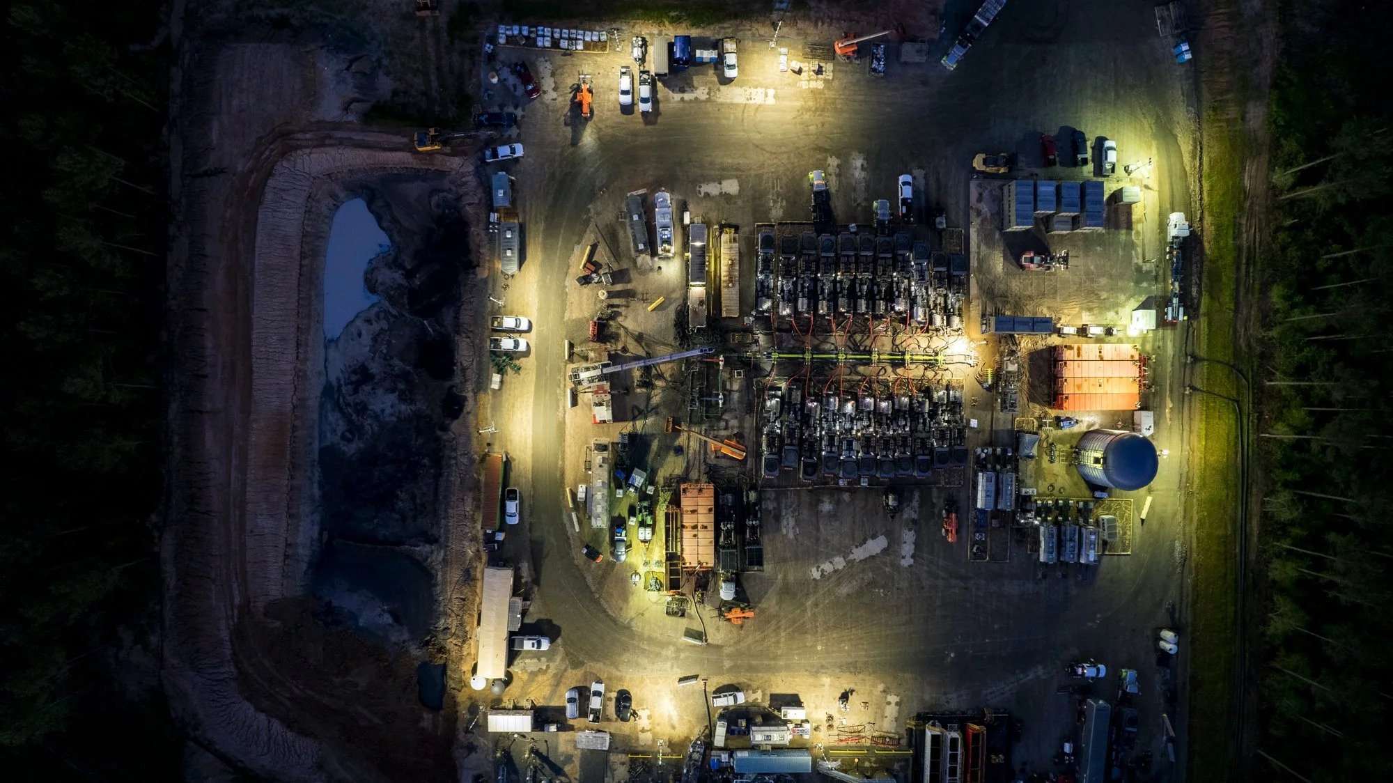 An aerial view of an industrial facility at night, with illuminated construction equipment, parked vehicles, and machinery, surrounded by trees and dirt paths.