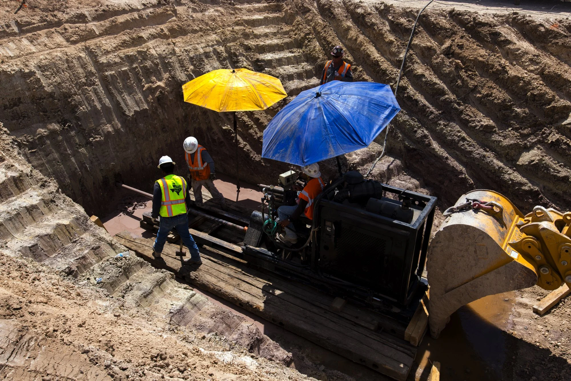 Construction workers excavating a trench with heavy machinery and shovels, some workers using umbrellas and wearing safety gear such as helmets and vests.