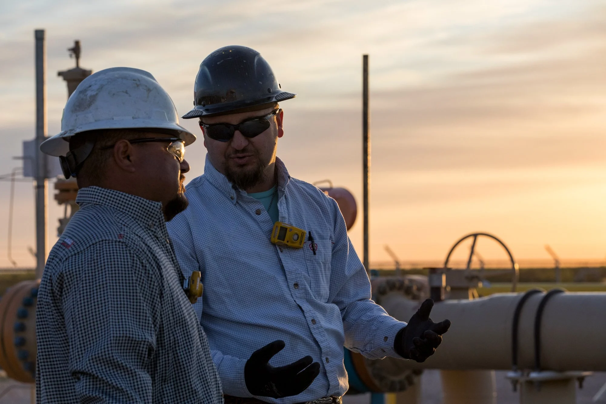 Two male workers in safety helmets and gloves standing near industrial pipes, having a conversation outdoors during sunset.