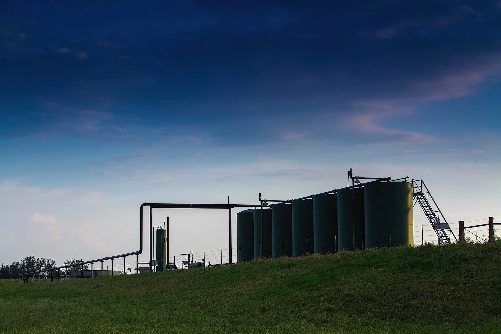 Industrial storage tanks surrounded by pipelines on a grassy field with a blue sky and clouds above.